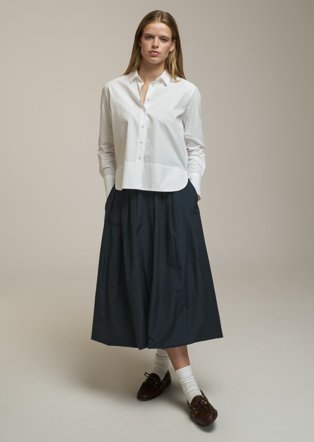 A woman stands against a plain warm beige studio background wearing a white long-sleeve button-front shirt, a dark navy midi skirt, white socks, and dark brown loafers.