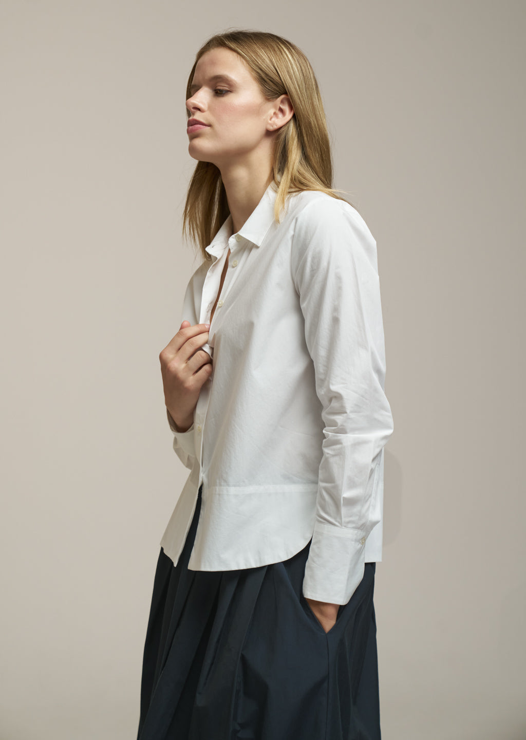A woman wearing a white long-sleeve button-up shirt and dark navy trousers, standing against a plain beige studio background.