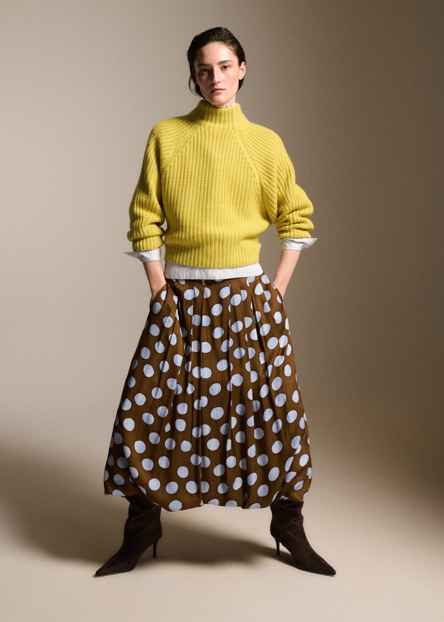 A model stands against a warm beige studio background wearing a bright yellow ribbed turtleneck sweater over a white collared shirt, paired with a brown skirt featuring a white polka-dot pattern, and dark brown pointed-toe heeled boots.