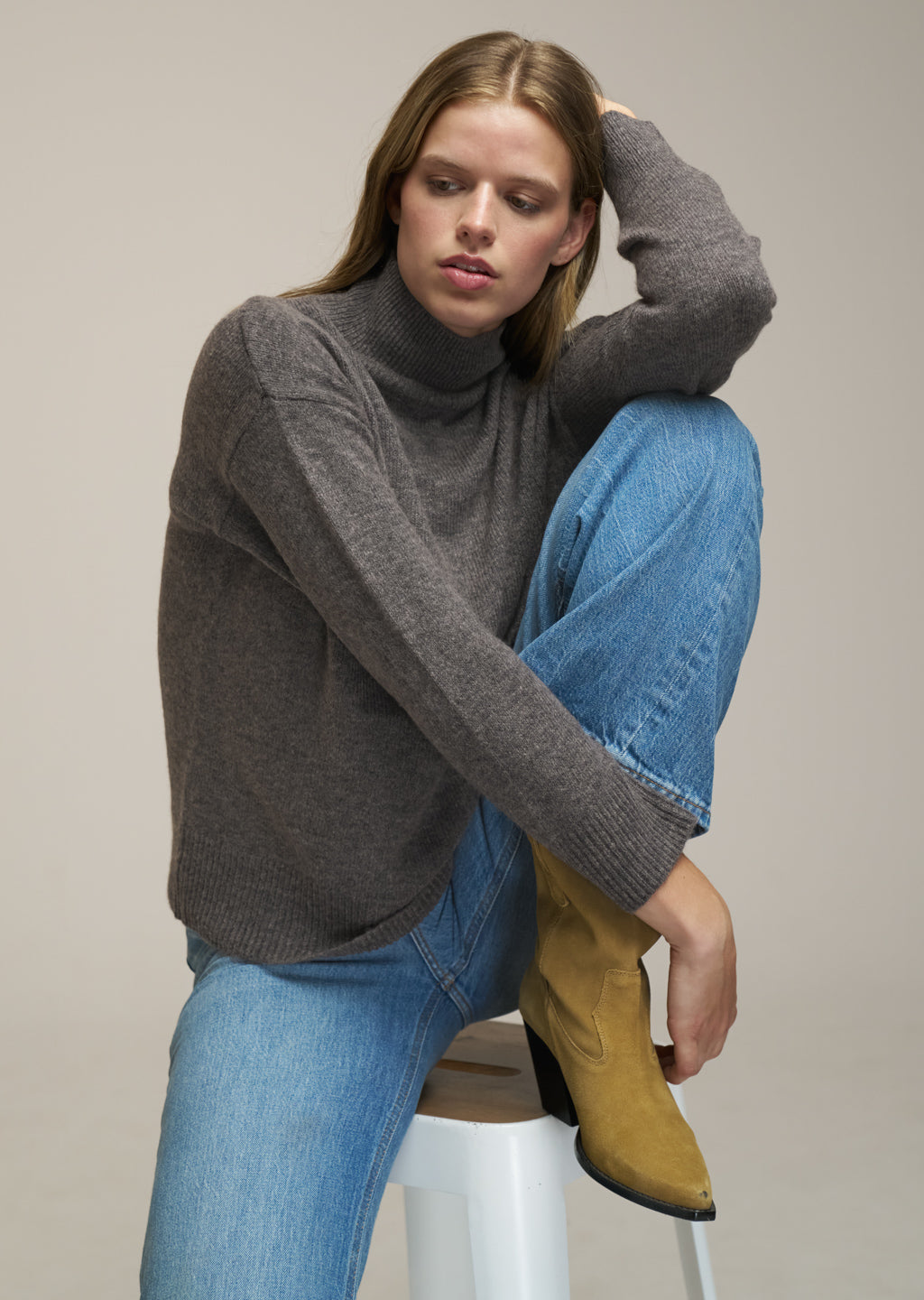 A woman models a dark gray turtleneck sweater, blue jeans, and a yellow ankle boot while sitting on a white stool against a neutral beige background.