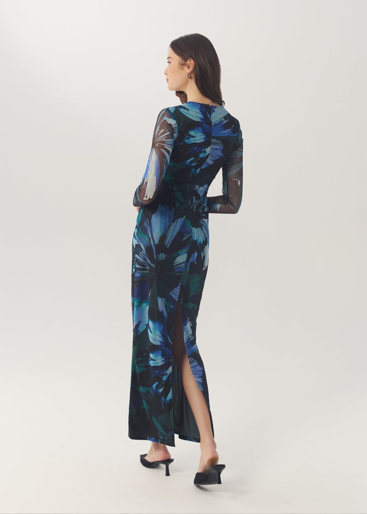 A woman modeling a long-sleeved maxi dress with a blue and black tropical leaf print, shown from a side-back angle against a light gray studio background.