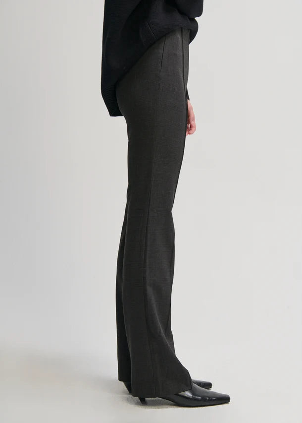 A side view of a person wearing dark charcoal gray flared trousers, a black top, and black pointed-toe flats against a light gray studio background.