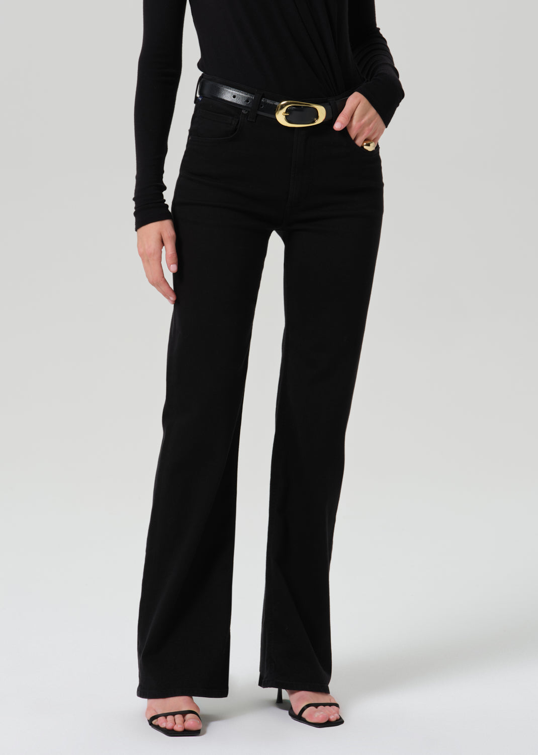 A model wearing black high-waisted flared jeans with a black leather belt featuring a gold oval buckle, paired with a black long-sleeve top and black sandals, against a light gray studio background.