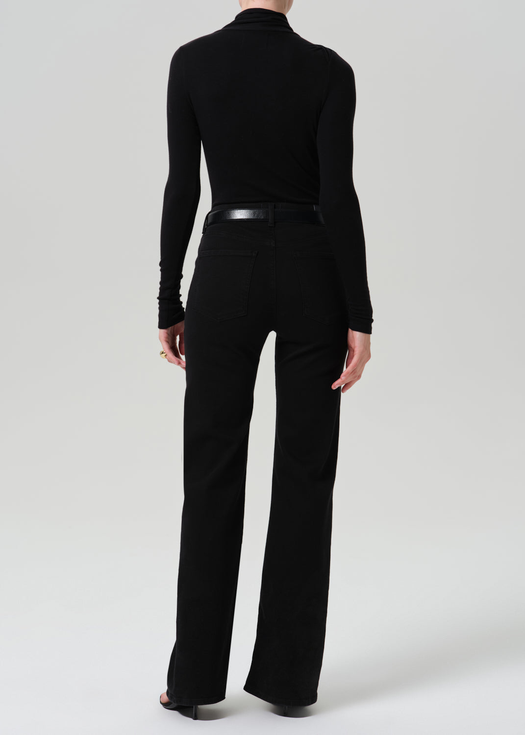A model shown from the back wearing a black long-sleeve mock neck top and black wide-leg trousers, paired with a black belt, against a light gray studio background.
