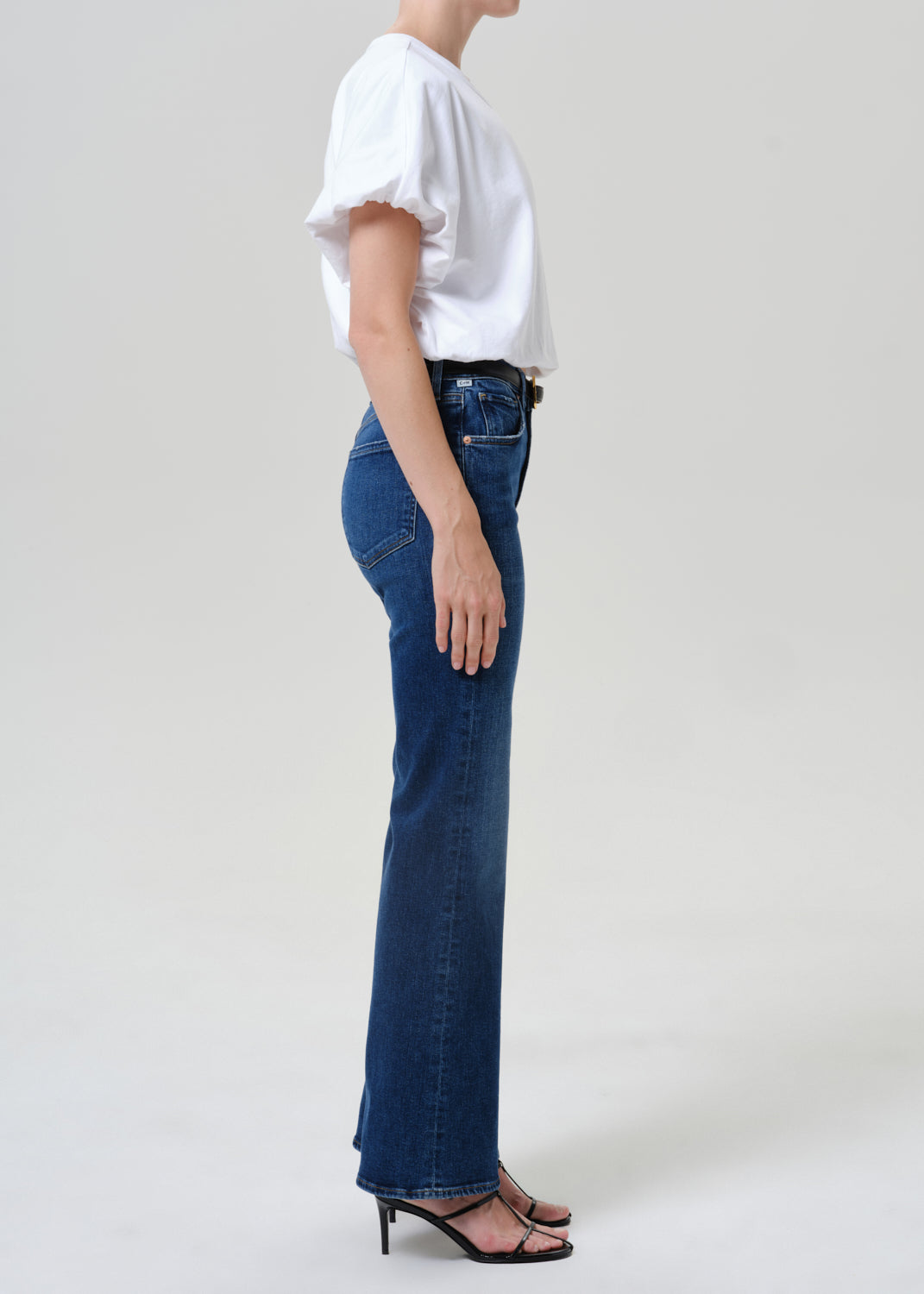 A side-profile view of a model wearing dark blue flare jeans, a white short-sleeve blouse, and black strappy heels against a light gray studio background.