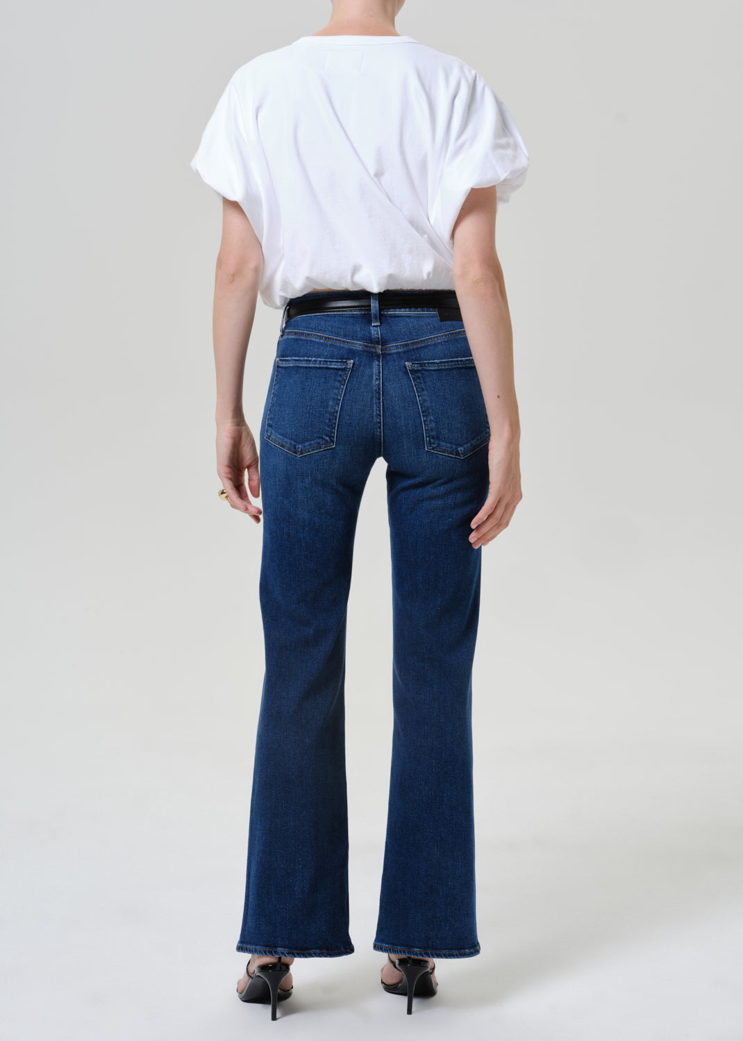 A model shown from the back wearing dark blue flare jeans, a white t-shirt, and black heels against a light gray studio background.