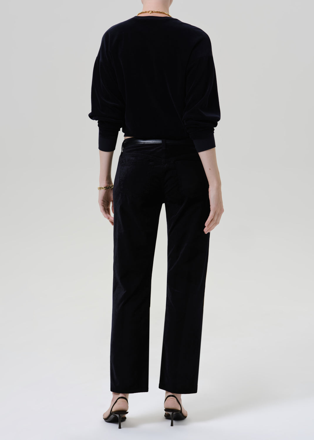 A model shown from the back wearing a black velvet long-sleeve top and matching black velvet wide-leg trousers, paired with black strappy heels against a light gray studio background.