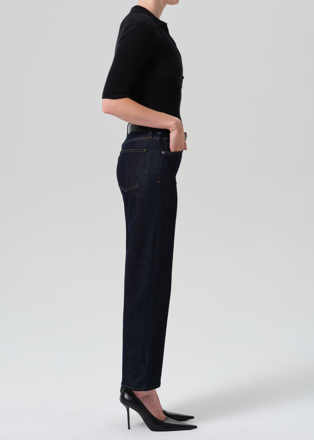 A side-profile studio shot of a model wearing dark wash straight-leg jeans, a black short-sleeve top, and black pointed-toe stiletto heels against a light gray background.