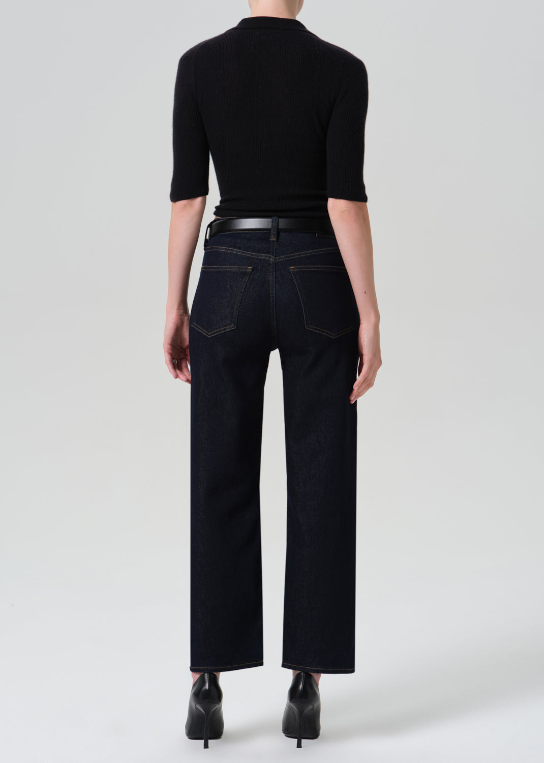 Rear view of a model wearing dark wash wide-leg jeans with a black belt, a black short-sleeve top, and black pointed-toe heels against a light gray studio background.