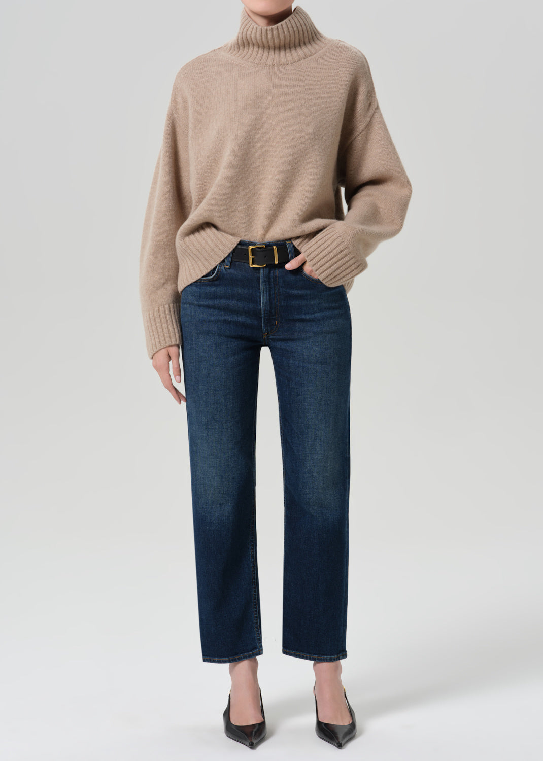 A model wearing a beige ribbed turtleneck sweater, dark blue straight-leg jeans with a gold-tone belt, and black pointed-toe heels, photographed against a light gray studio background.