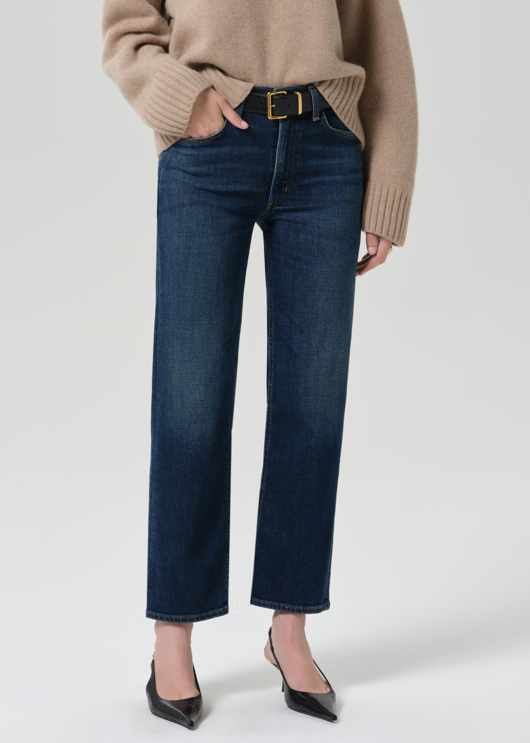 A model wearing dark blue straight-leg jeans with a gold-tone belt, styled with a beige sweater and black pointed-toe heels against a light gray studio background.