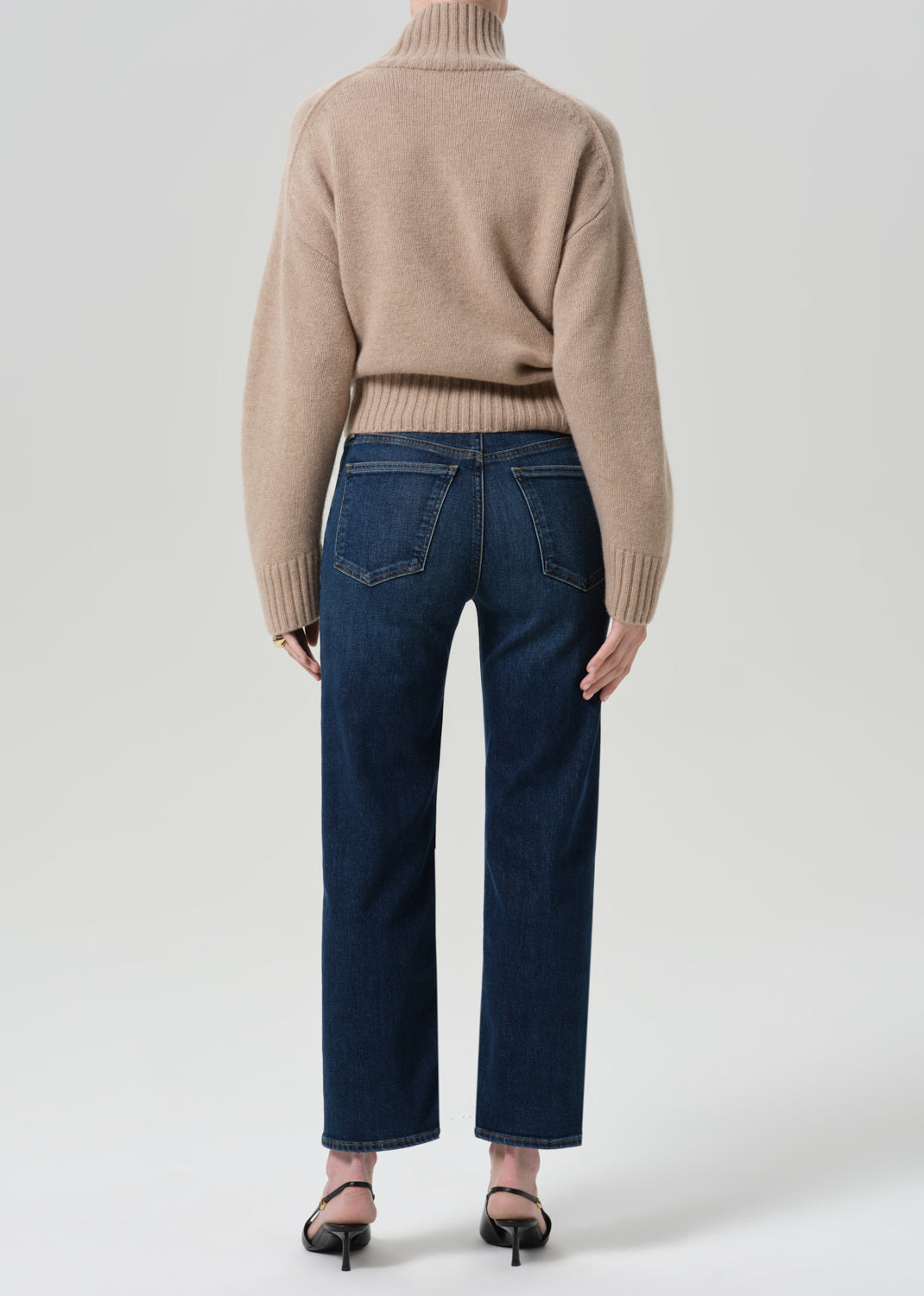 A model shown from the back wearing a beige ribbed turtleneck sweater and dark blue straight-leg jeans, paired with black pointed-toe heels, against a light gray studio background.