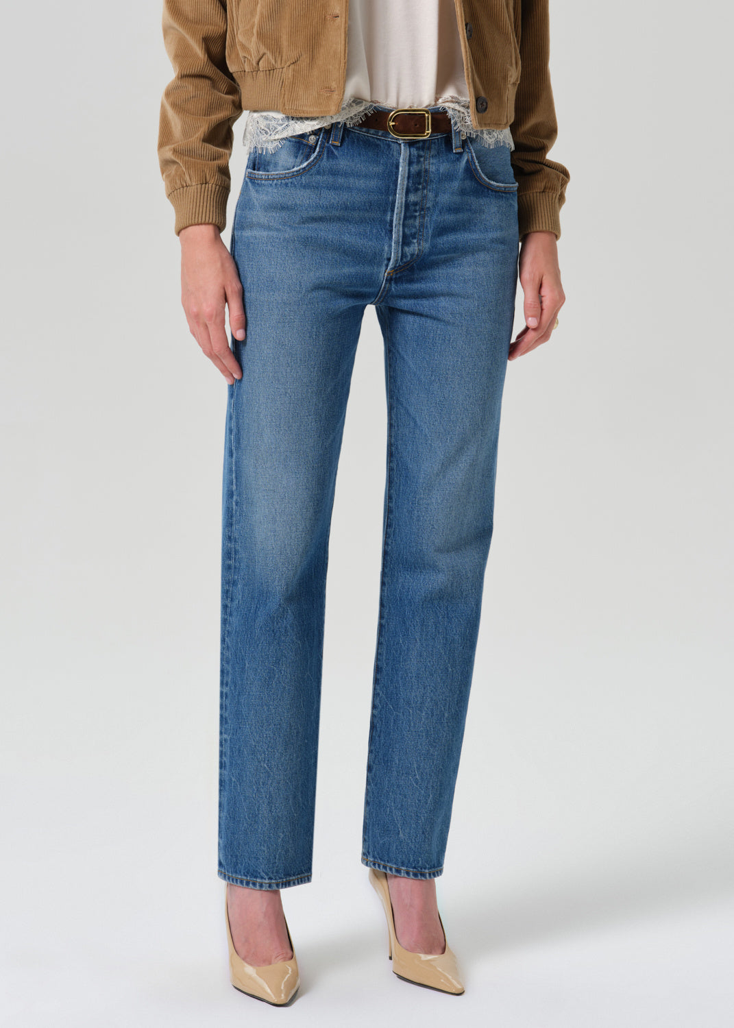 Front view of a model wearing blue straight-leg jeans with a raw, frayed hem, paired with a brown suede jacket and beige heels, against a light gray studio background.