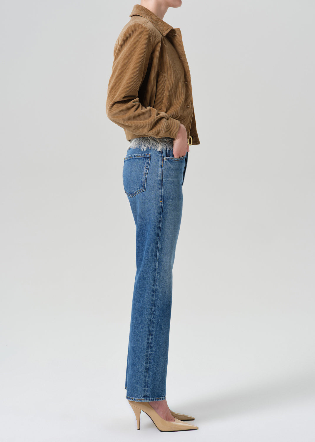 A side-profile view of a model wearing blue straight-leg jeans, a brown jacket, and beige heels against a light gray studio background.