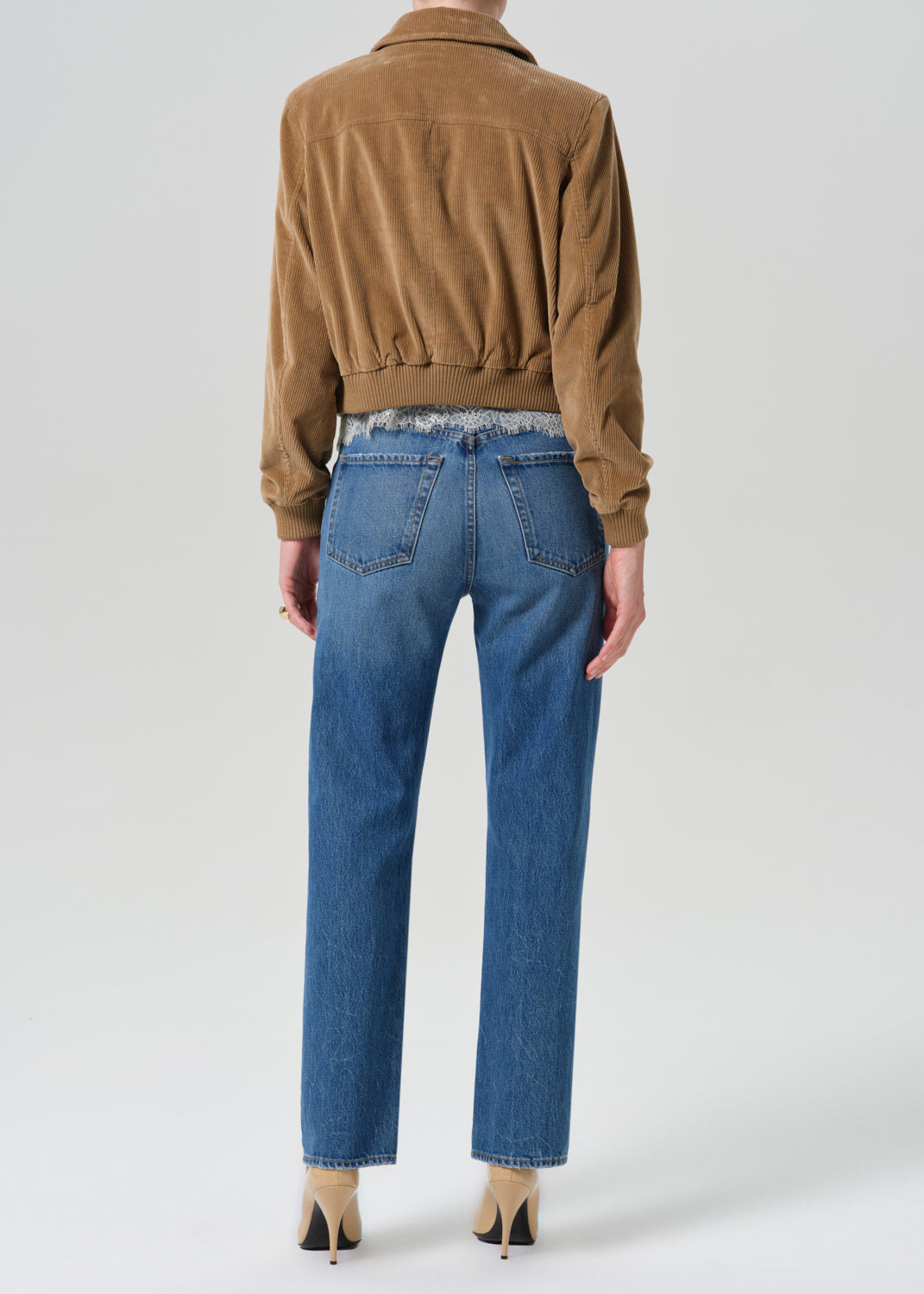 Rear view of a model wearing blue straight-leg jeans with a raw, frayed hem, paired with a brown suede jacket and beige heels, against a light gray studio background.
