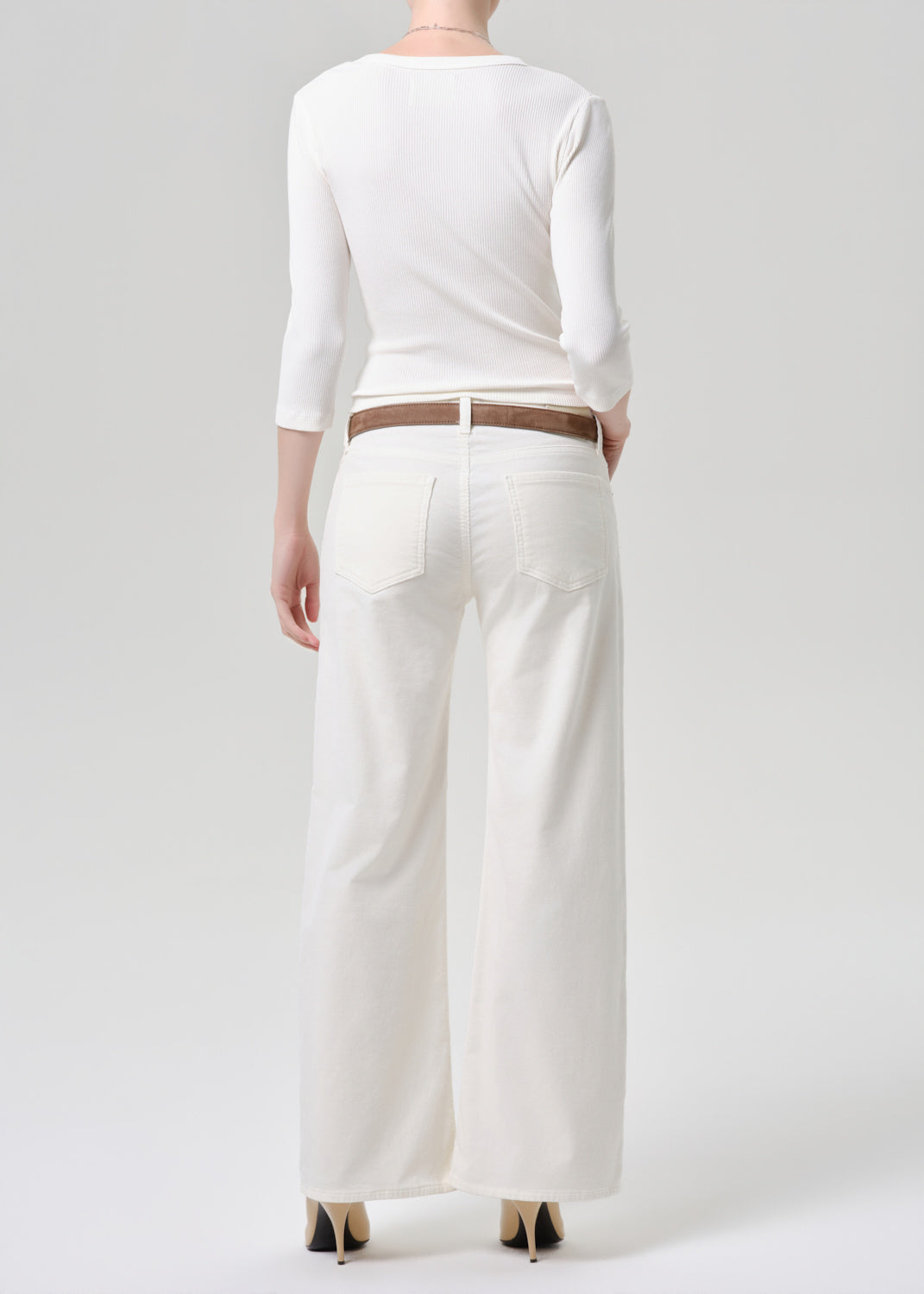 A model shown from the back wearing a white three-quarter sleeve top, white wide-leg trousers, a thin brown belt, and black pointed-toe heels against a light gray studio background.