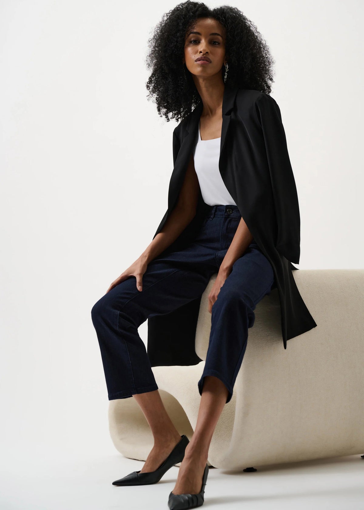Woman in black coat and navy pants sitting on a white couch against a white background