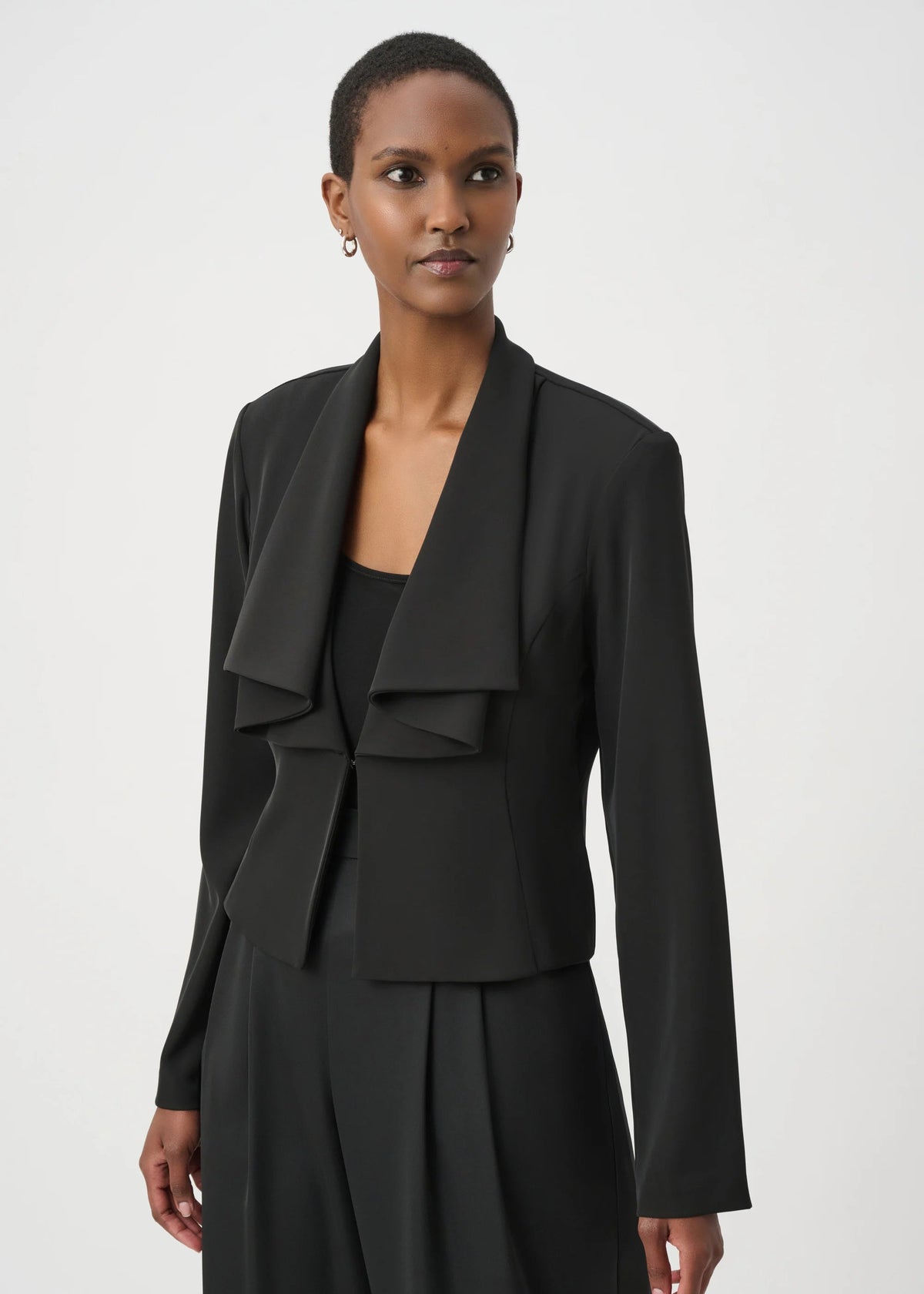A model wearing a black draped ruffle jacket with long sleeves, paired with a matching black skirt, photographed against a light gray studio background.