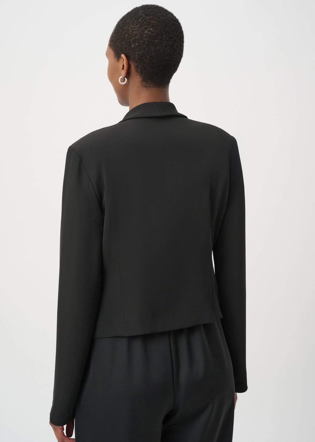 Rear view of a model wearing a black long-sleeve cropped blazer with matching black trousers against a light gray studio background.
