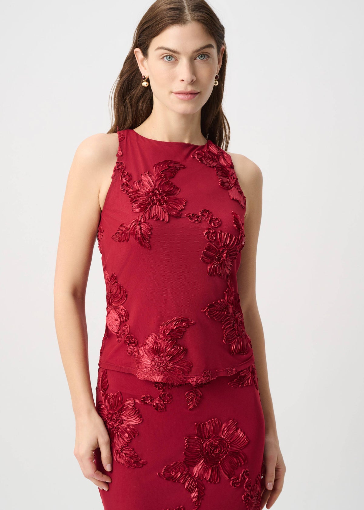 A woman modeling a sleeveless red dress with large floral applique and sequined detailing against a light gray studio background.