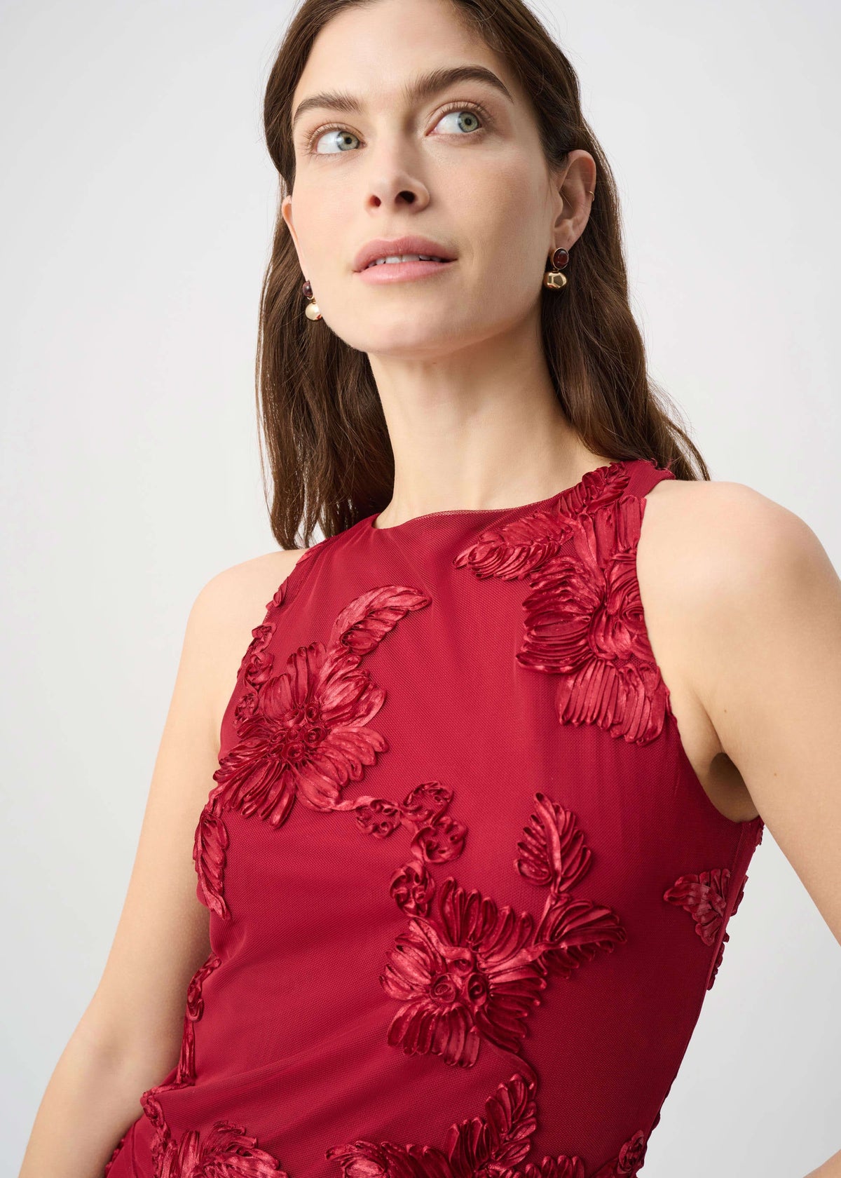 A woman wearing a sleeveless deep red dress with 3D floral applique, photographed against a light gray studio background.
