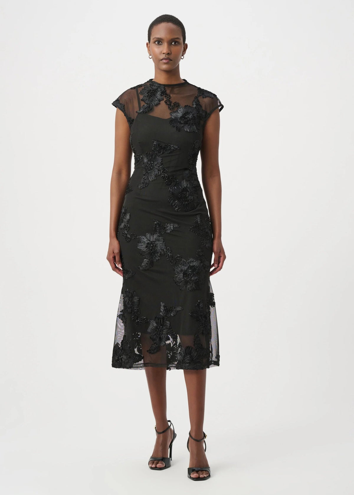A model wearing a black floral lace midi dress with sheer cap sleeves and a high neckline, paired with black strappy heels, against a plain white studio background.