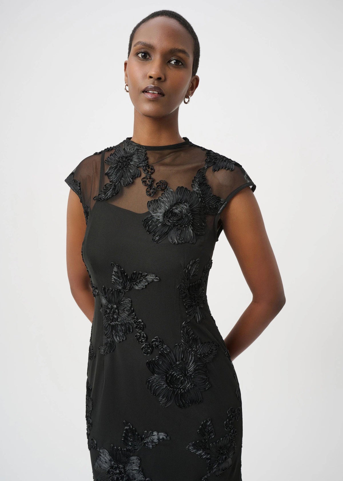 A woman modeling a black sheer mesh and floral lace dress against a light gray studio background.