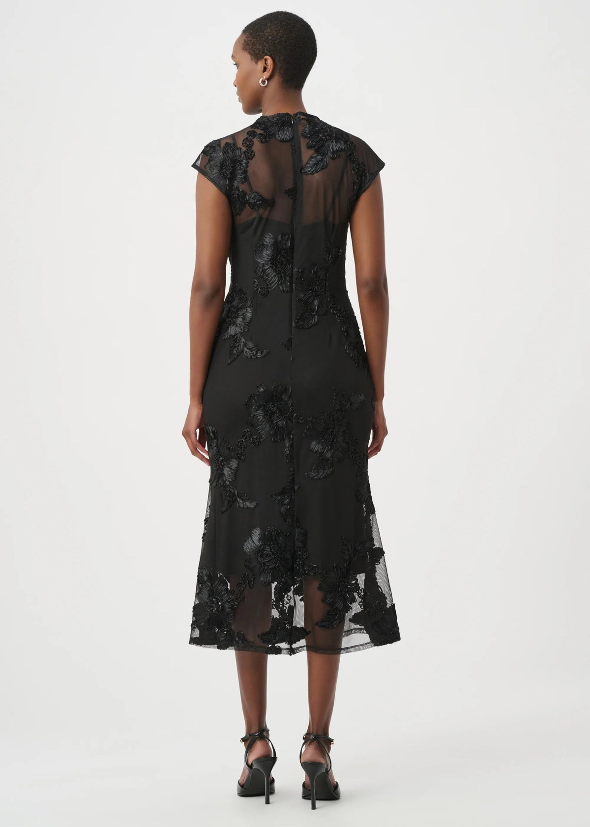 A woman viewed from the back wearing a black sheer lace midi dress with cap sleeves and black high heels, photographed against a plain light gray studio background.