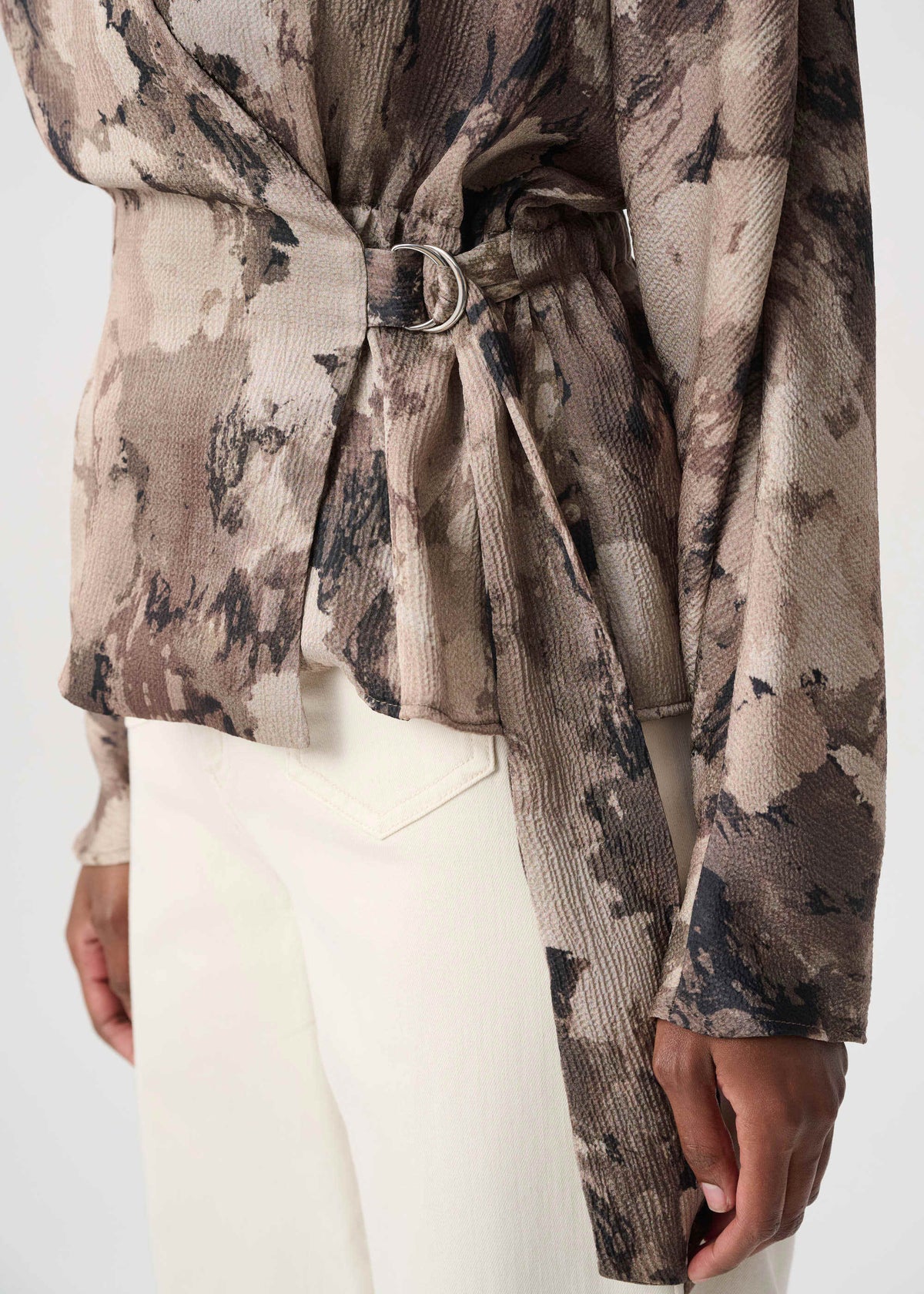 A close-up of a model wearing a long-sleeve belted top with a textured crinkle fabric and an abstract camouflage-like print in beige, brown, and black, paired with white trousers.