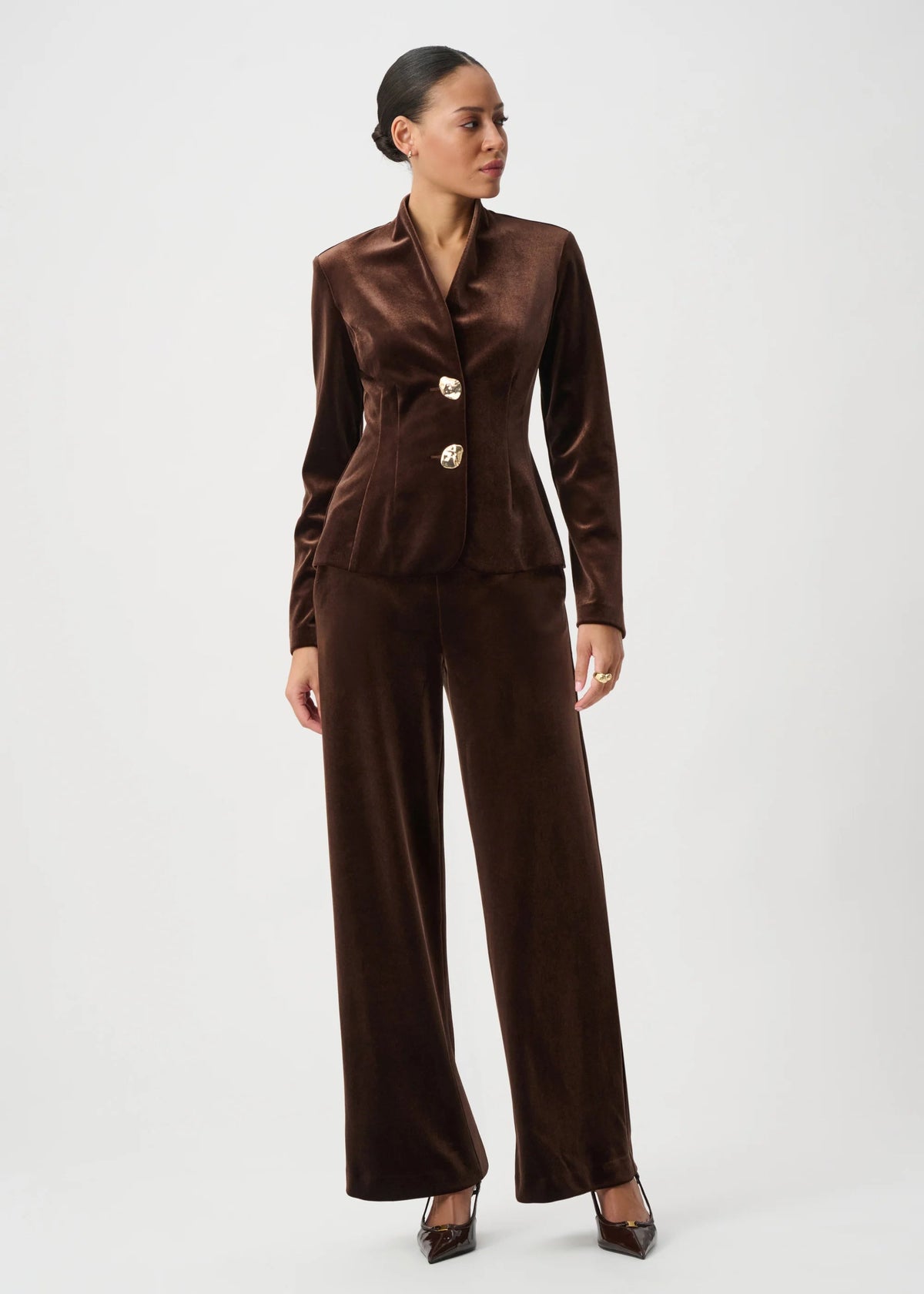 A model wearing a dark brown velvet two-piece suit with a structured blazer and wide-leg trousers, styled with dark brown pointed-toe heels against a light gray studio background.