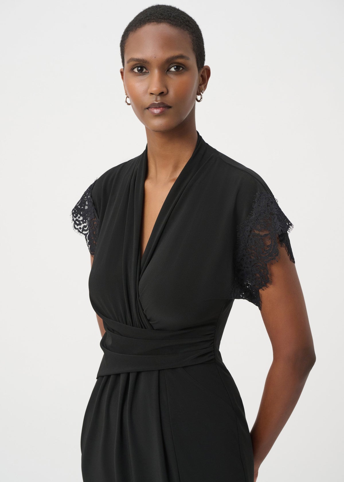 A woman modeling a black wrap-style dress with short flutter sleeves featuring black lace trim, photographed against a plain light gray studio background.