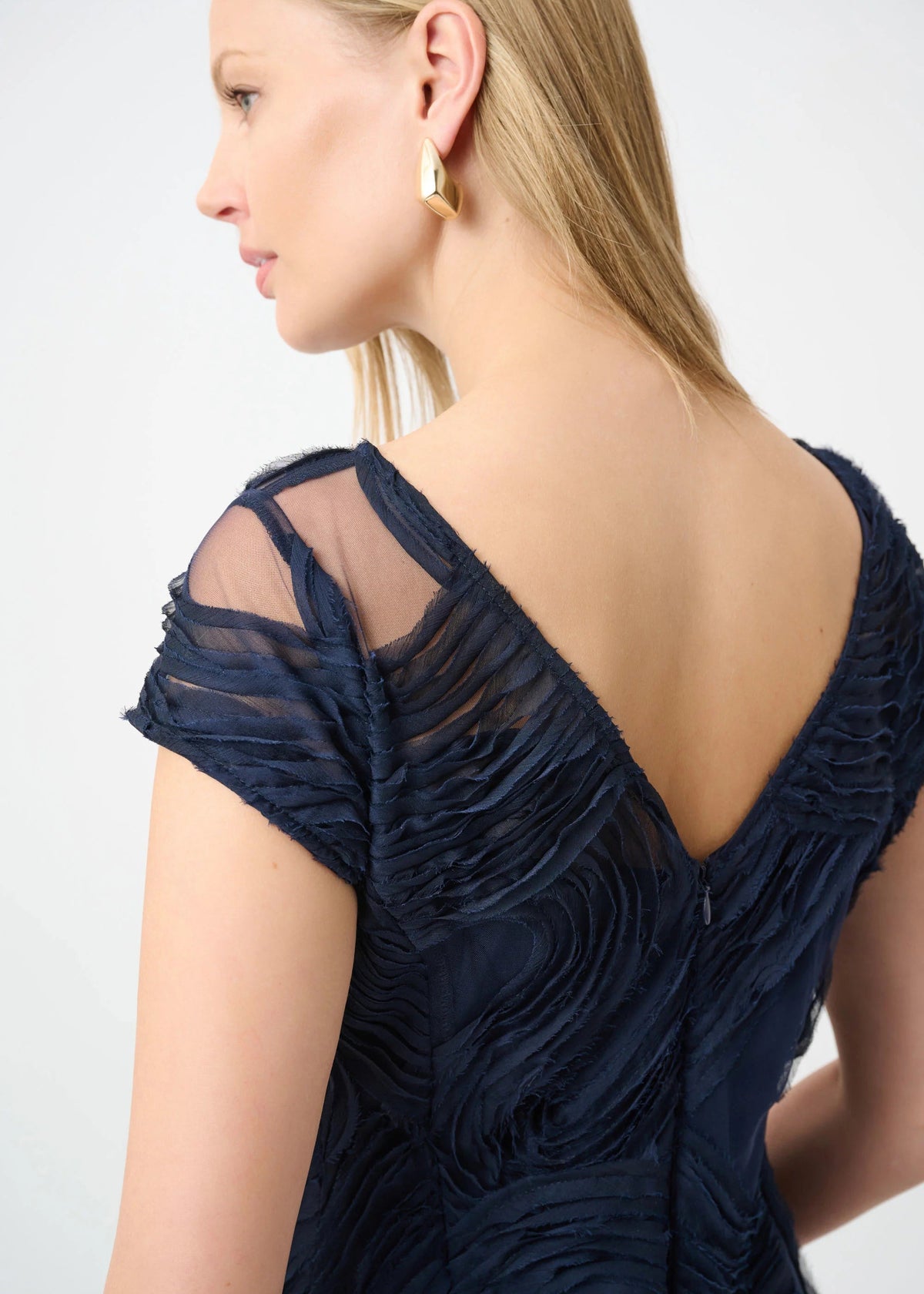 A woman shown from the back wearing a dark navy blue textured dress with sheer mesh shoulder panels and a deep V-neckline, paired with a gold earring, against a light gray studio background.