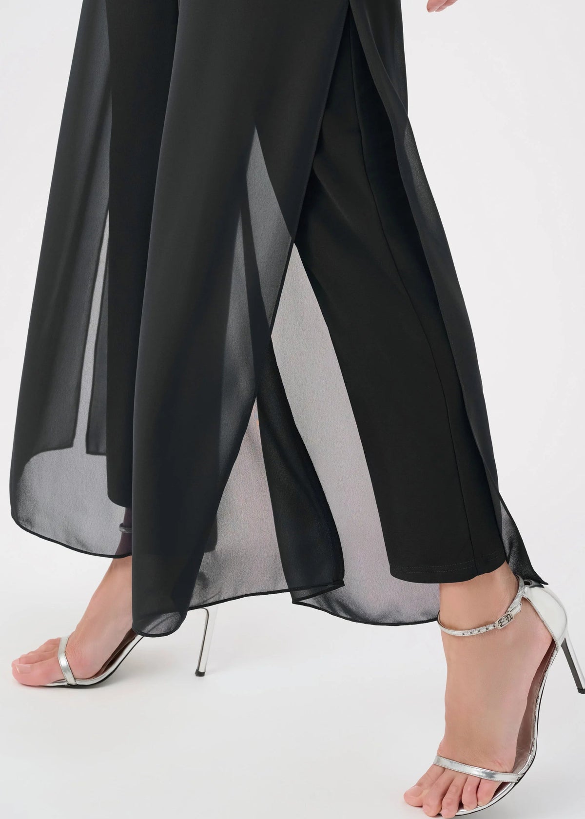 Black wide-leg draped pants with sheer side panels and a high slit, shown on a model wearing silver strappy high-heeled sandals against a light gray studio background.