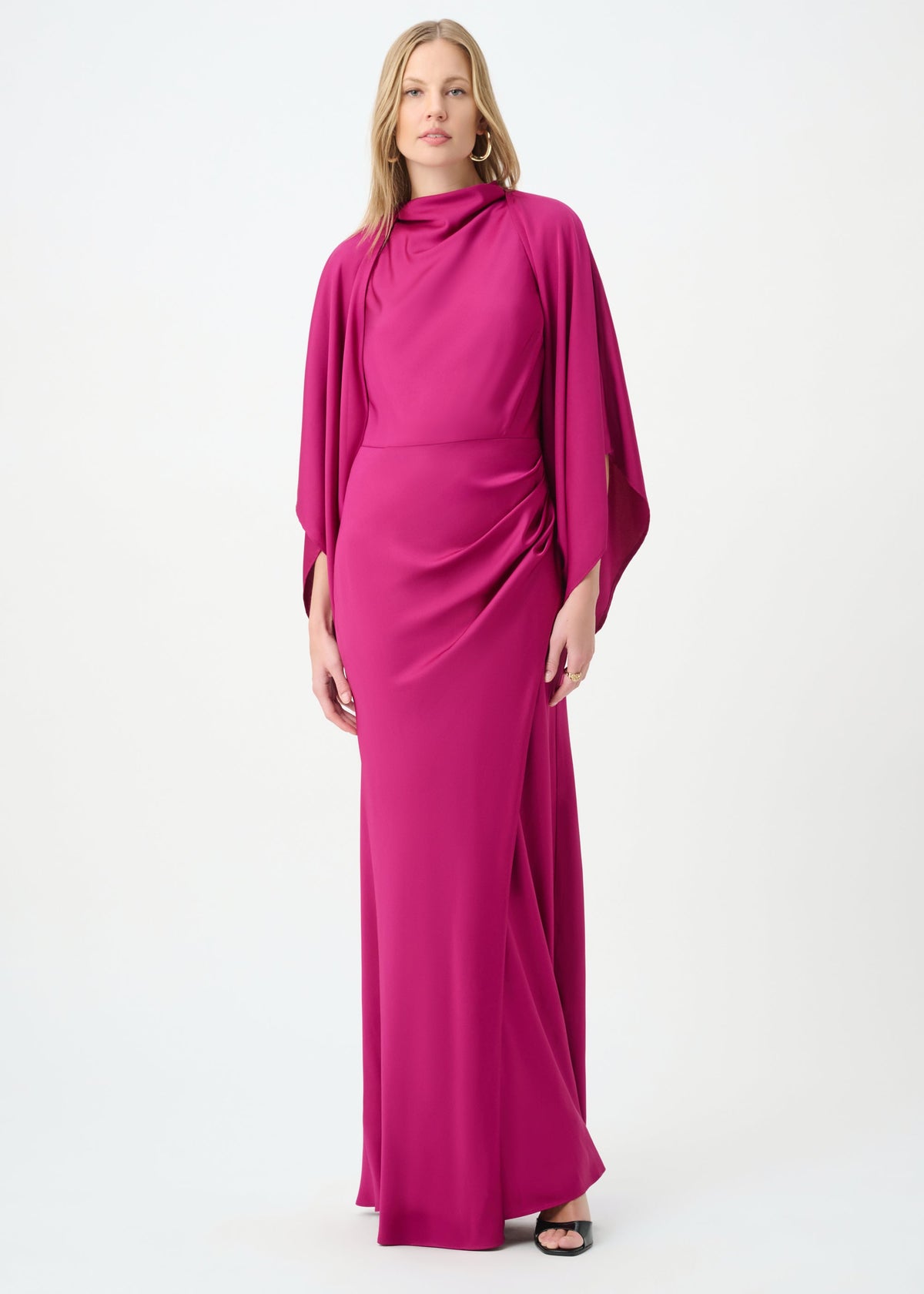 A woman modeling a floor-length magenta dress with a draped, high-neck silhouette and long flowing sleeves, photographed against a plain white studio background.
