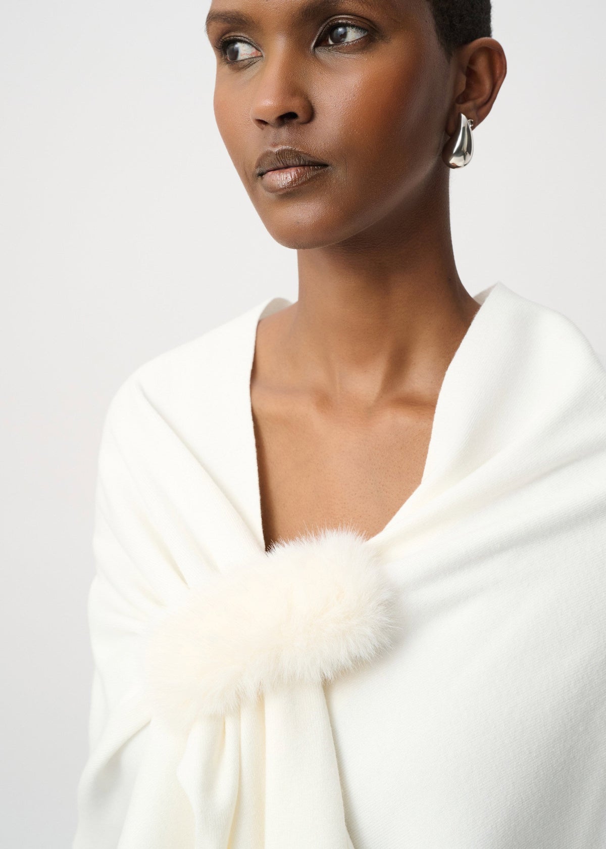 A close-up of a model wearing a white shawl with a fluffy faux-fur trim detail at the neckline, set against a plain light gray background.