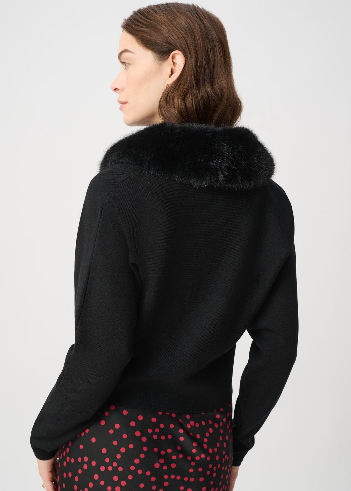 A woman shown from the back wearing a black long-sleeve top with a dark faux-fur collar, paired with a red and black polka-dot skirt, against a light gray studio background.