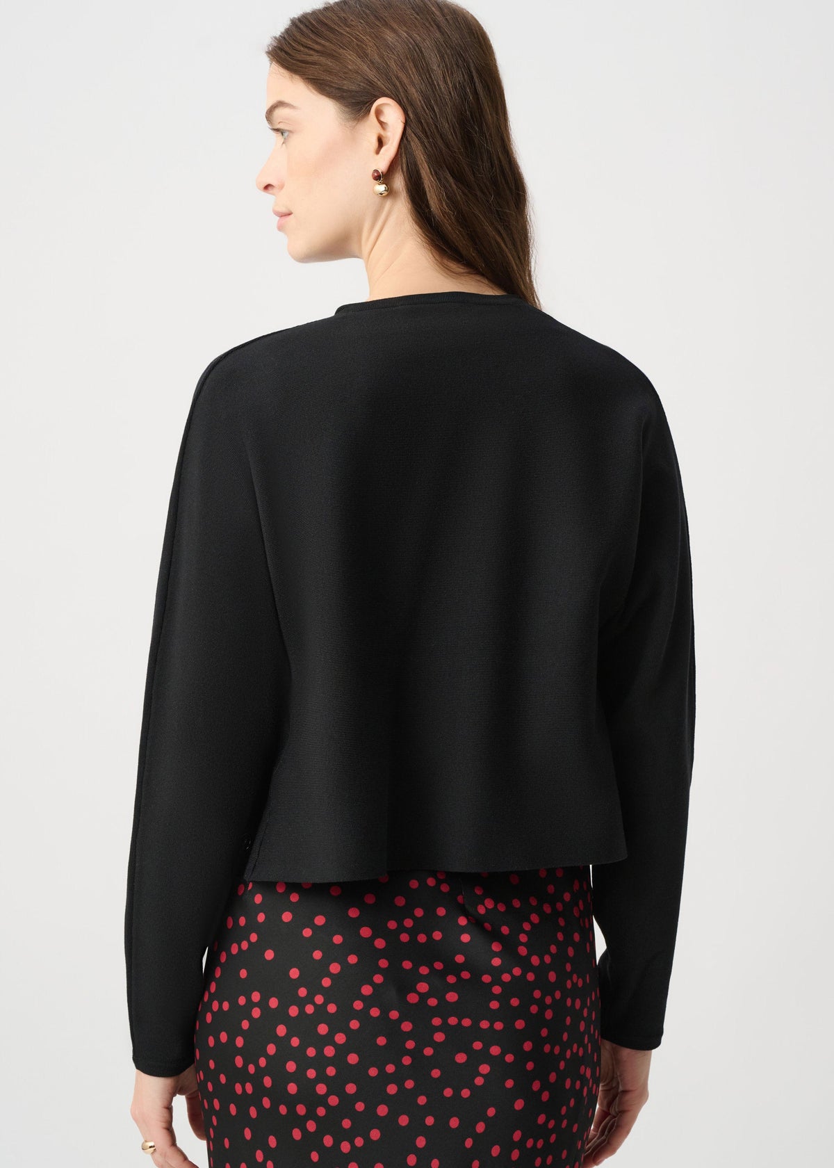 A woman shown from the back wearing a black long-sleeve cropped cardigan or jacket over a black skirt with red polka dots, against a light gray studio background.
