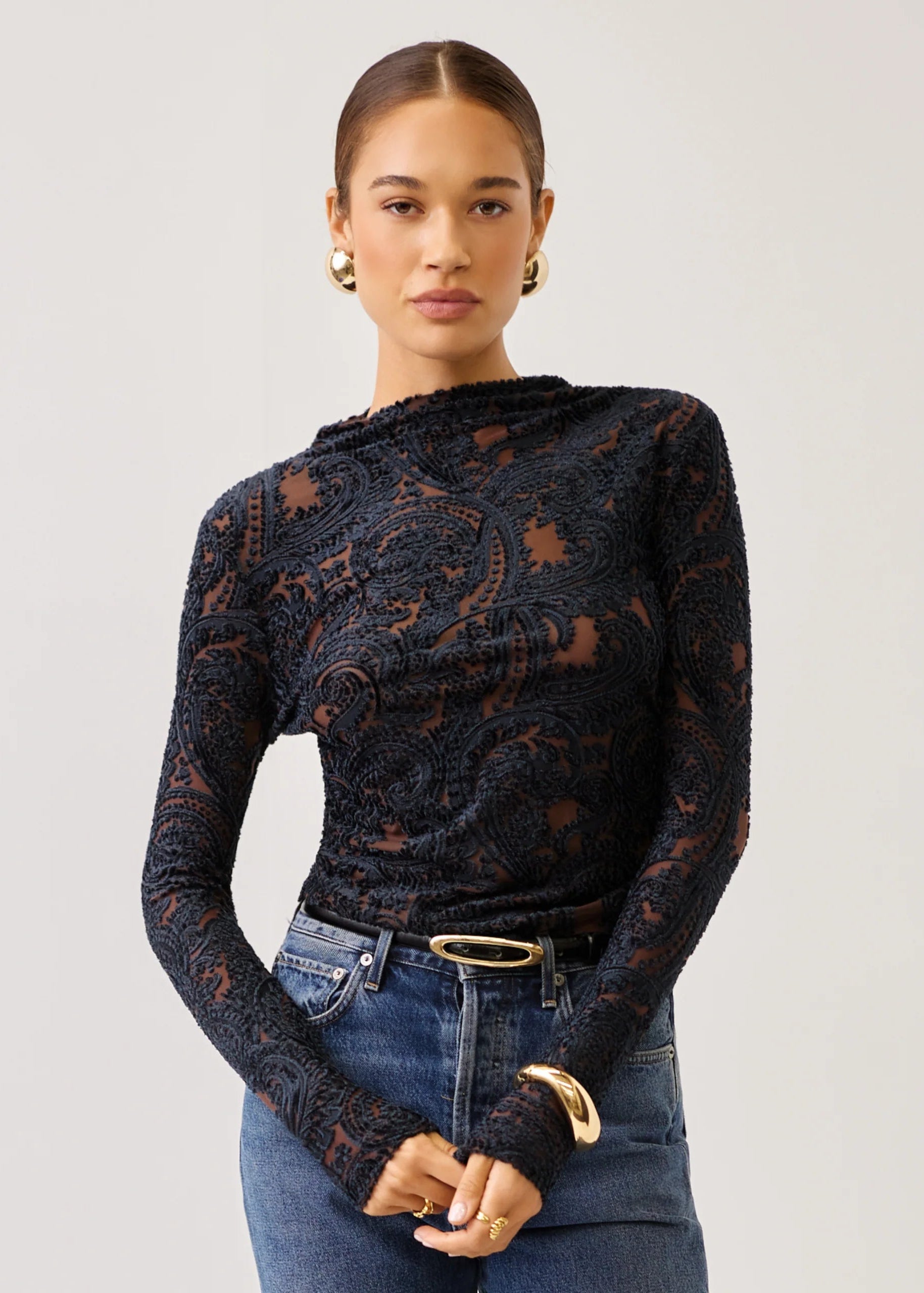 A woman modeling a black long-sleeve lace top with an intricate paisley pattern, paired with blue jeans and gold jewelry, against a plain light gray background.