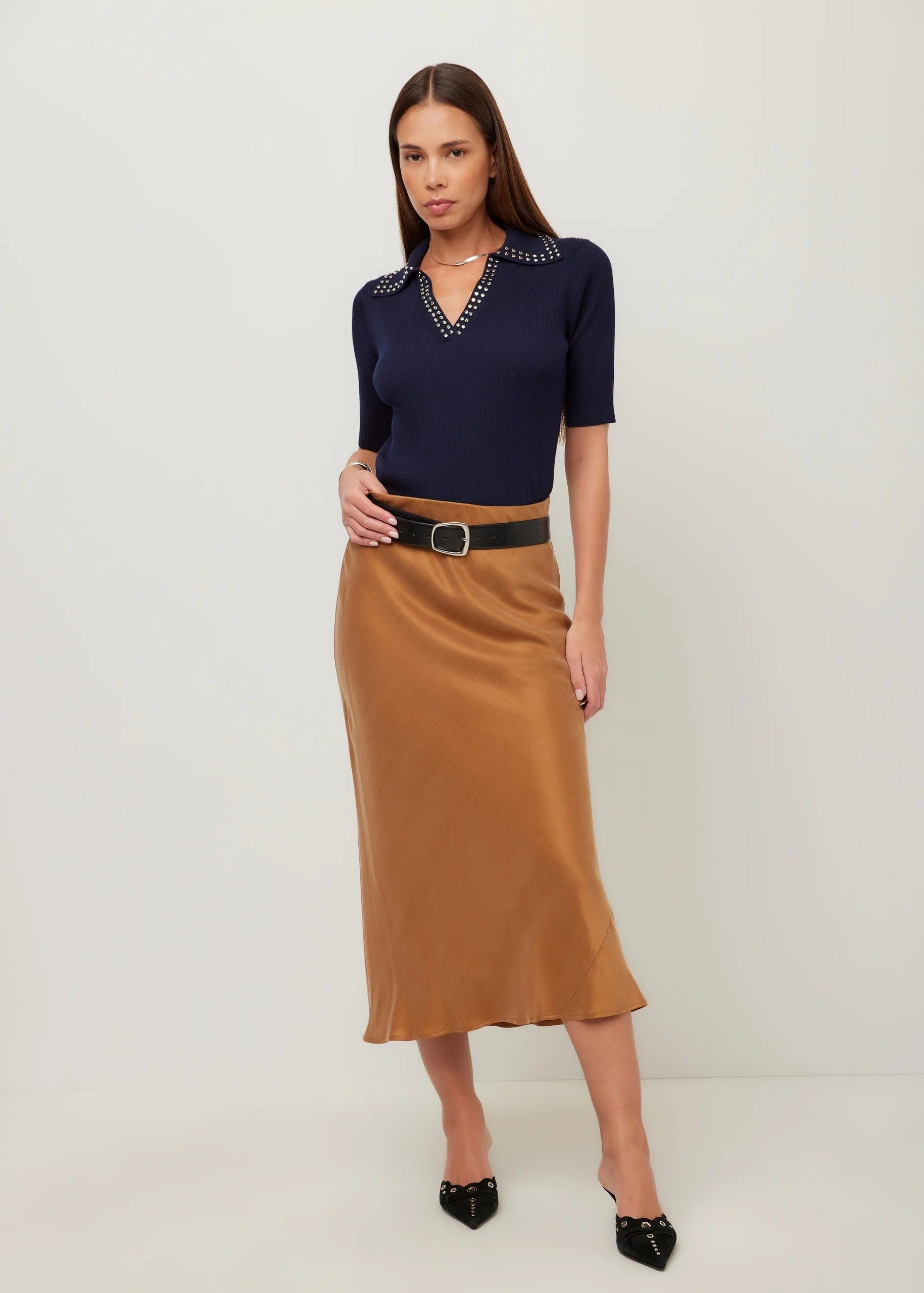 A woman modeling a navy blue short-sleeve knit top with silver embellished collar detailing, paired with a brown satin midi skirt and black pointed-toe heels, against a plain off-white studio background.