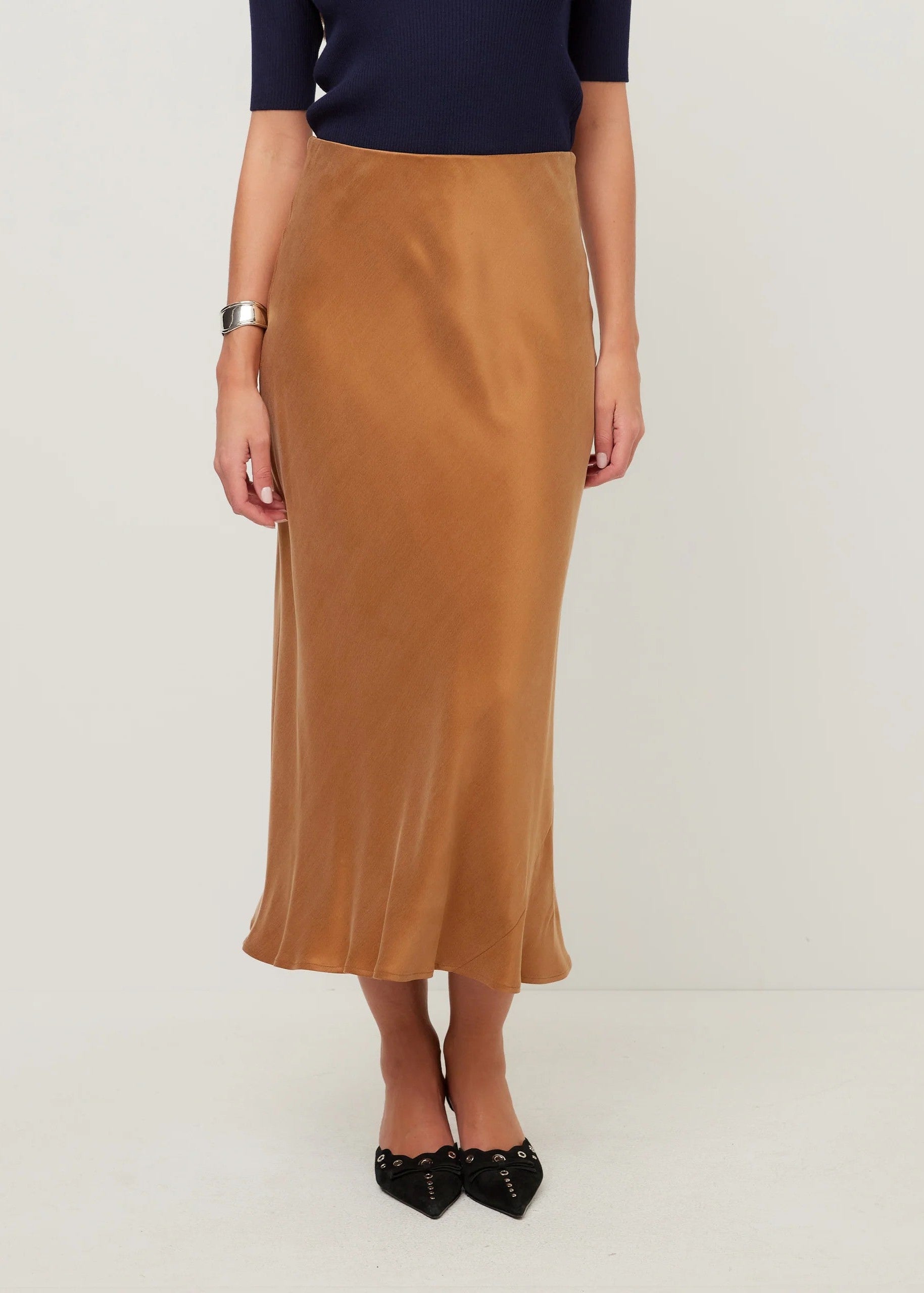 A woman wearing a satin-like camel brown midi skirt with a navy blue top and black pointed flats, standing against a plain light gray studio background.