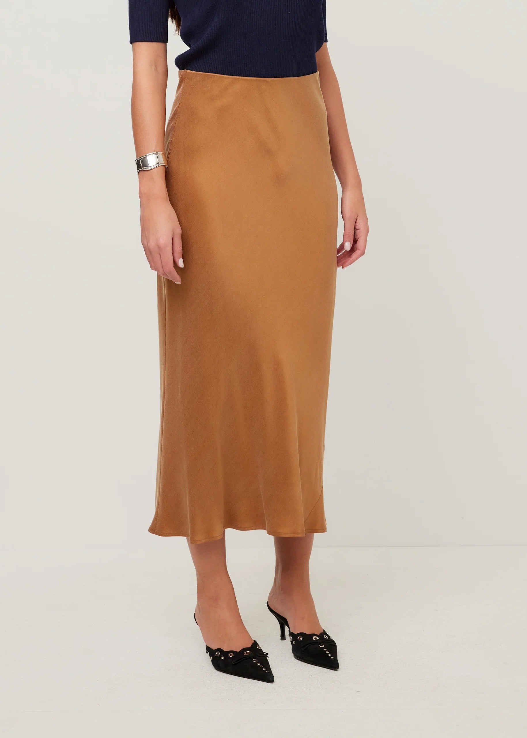A woman wearing a high-waisted, knee-length camel brown satin midi skirt, styled with a navy blue top and black pointed-toe heels with silver studs, against a plain light gray studio background.