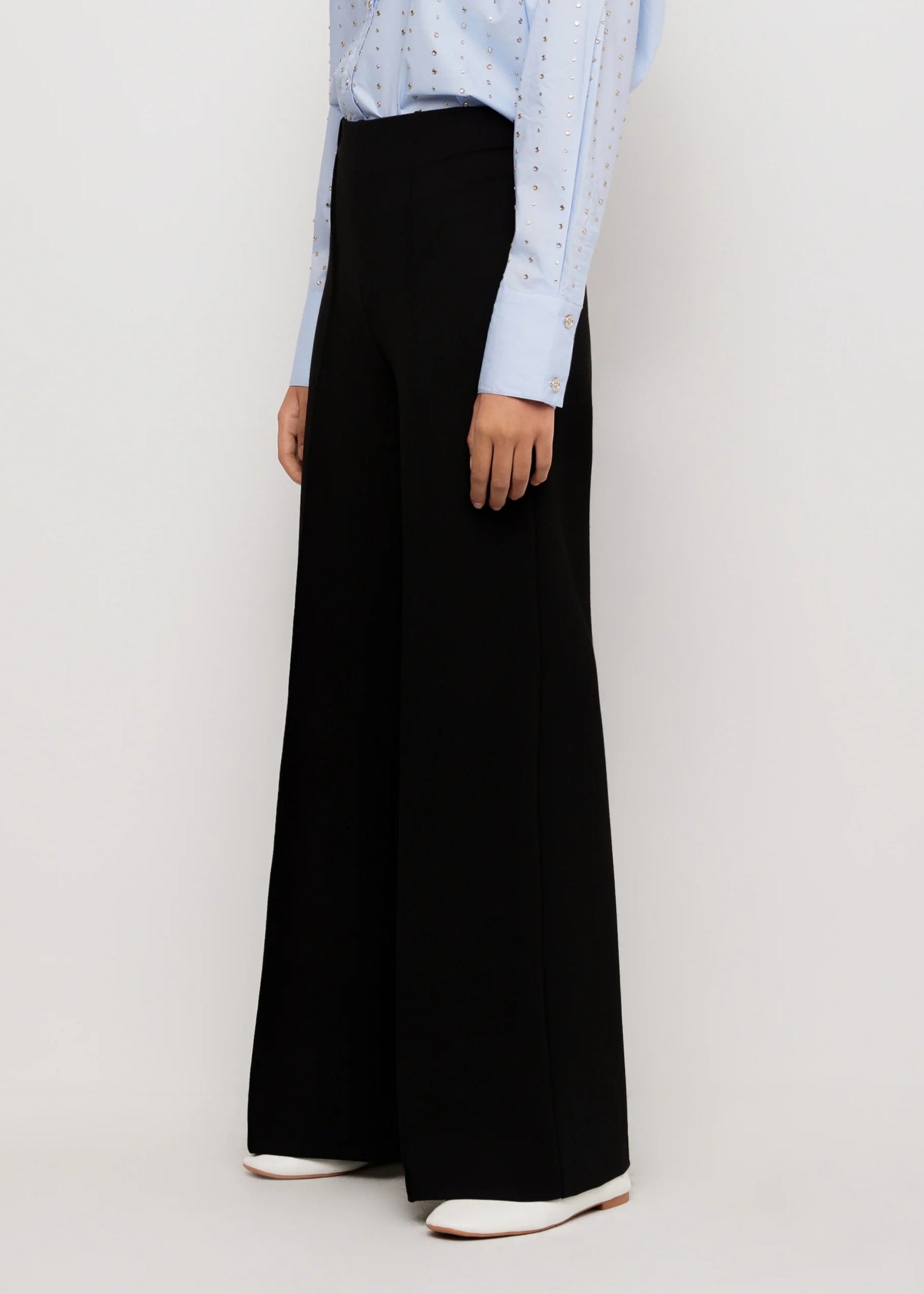 A model wearing high-waisted, wide-leg black trousers paired with a light blue patterned long-sleeve shirt and white pointed-toe flats, shown from the waist down against a light gray studio background.