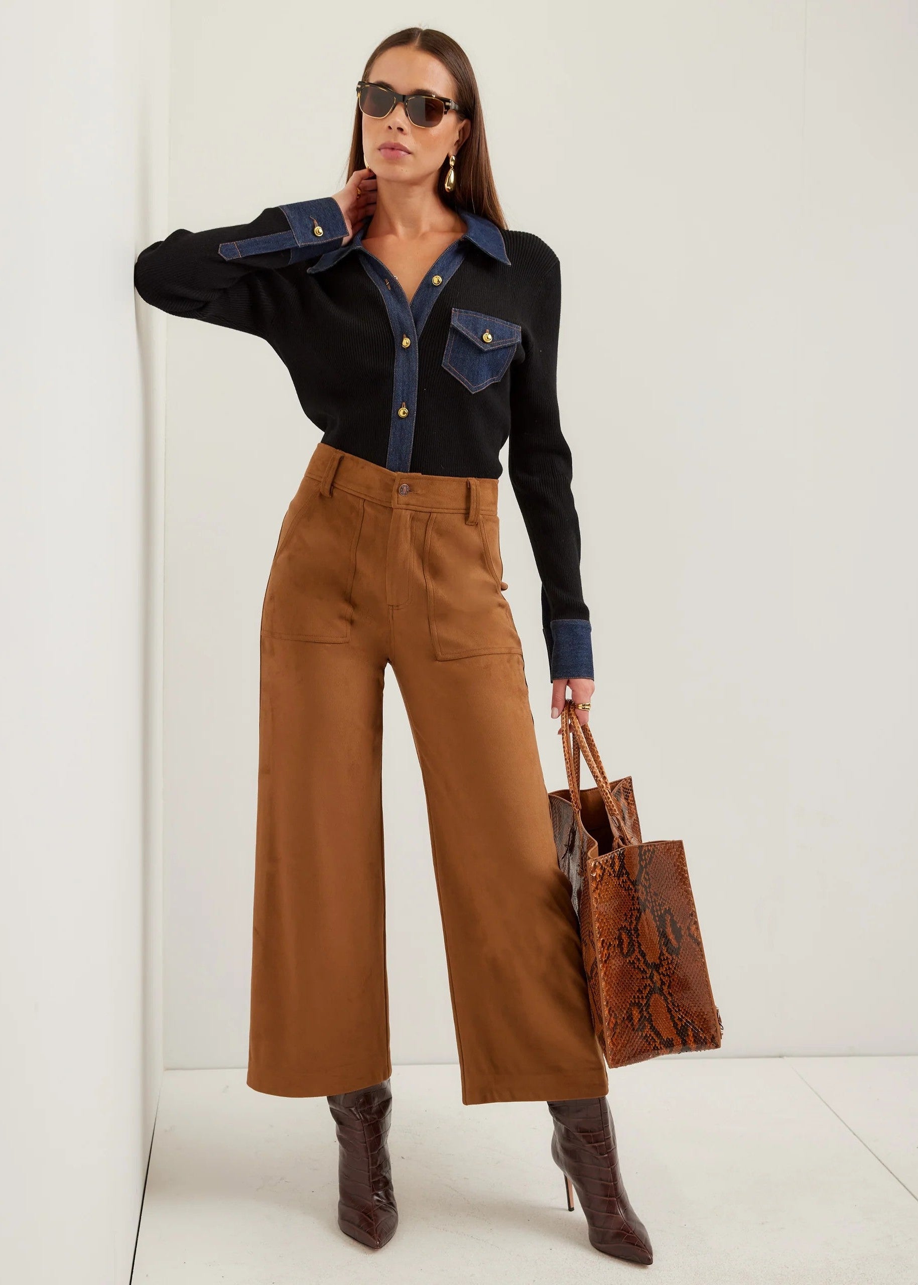 A model wearing a black and navy long-sleeve button-up shirt with denim-style chest pockets, paired with high-waisted brown wide-leg trousers and dark brown heeled boots, holding a brown snakeskin-patterned tote bag against a white studio background.