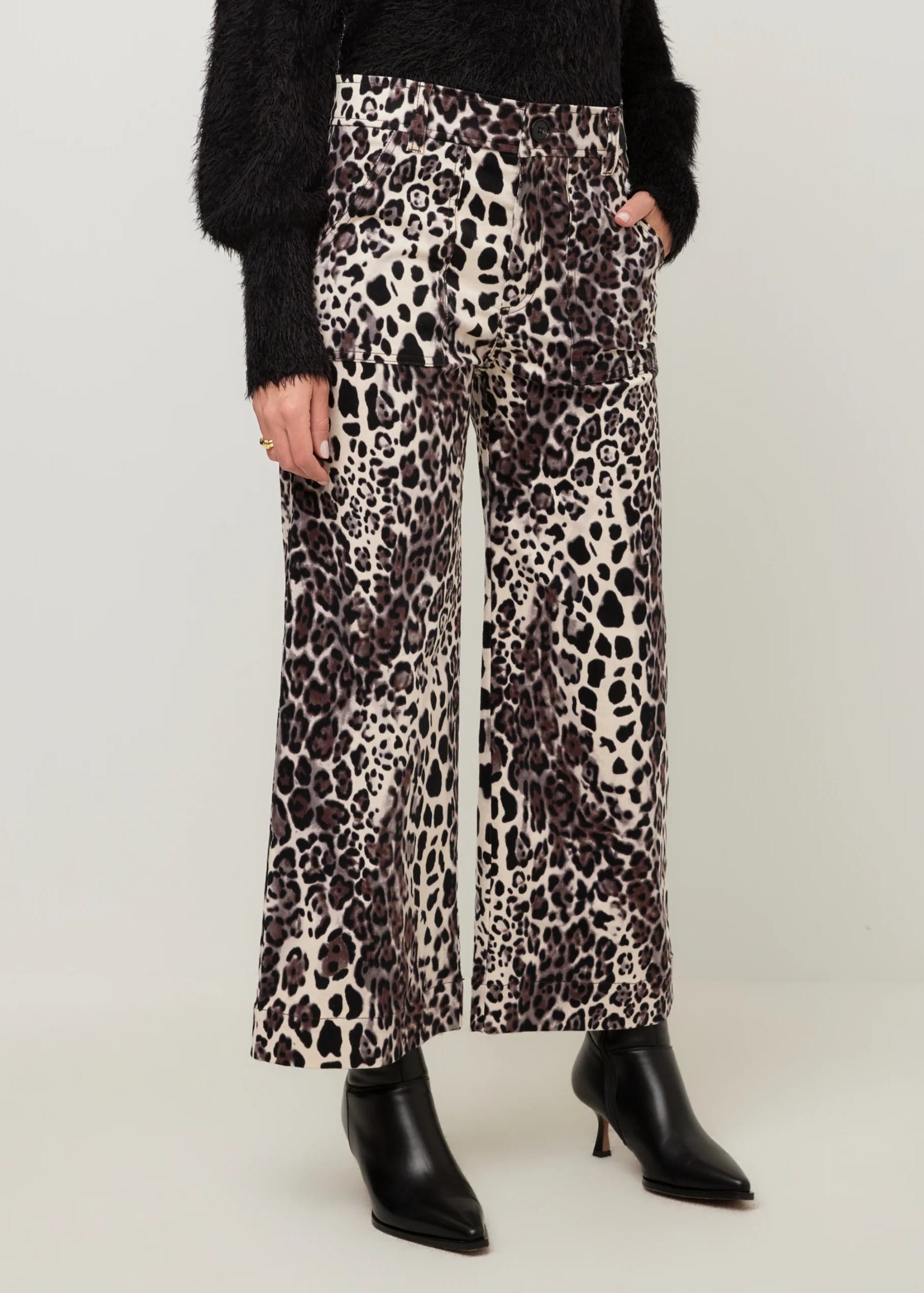 A model wearing high-waisted, wide-leg trousers with a black, brown, and beige leopard print, styled with a black fuzzy sweater and black pointed-toe heeled boots against a light gray studio background.