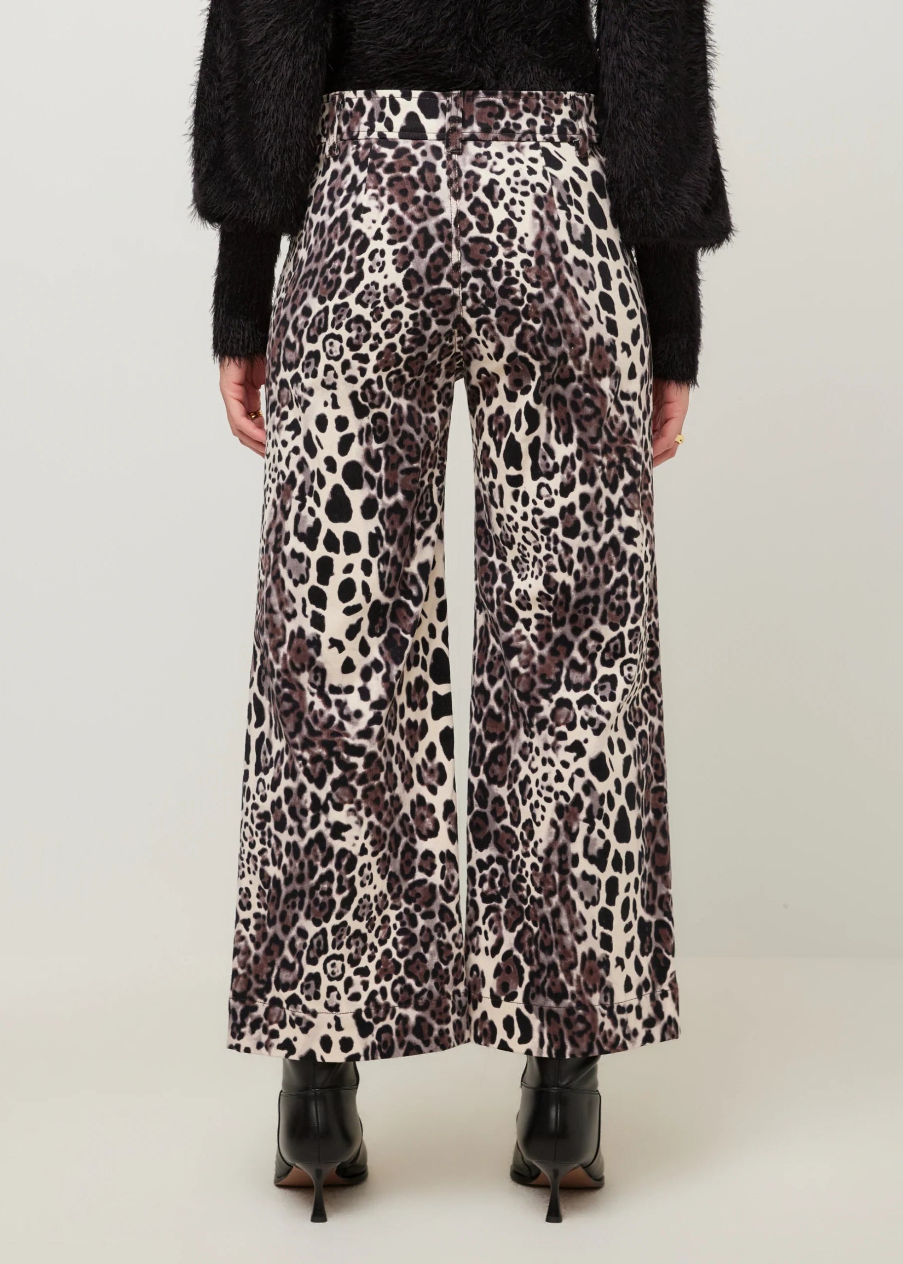 A person shown from the back wearing wide-leg leopard-print pants, a black fuzzy top, and black pointed-toe heels against a plain light gray studio background.