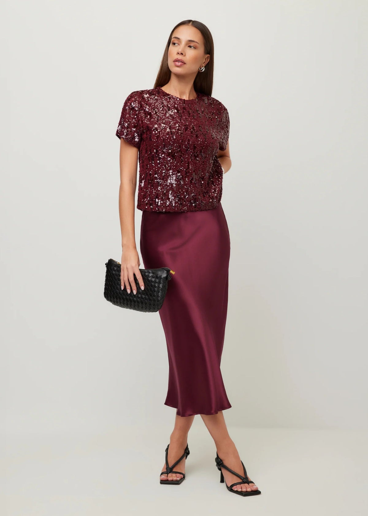 A woman models a burgundy sequined short-sleeve top paired with a matching satin midi skirt, holding a black woven clutch and wearing black strappy heels against a light gray studio background.
