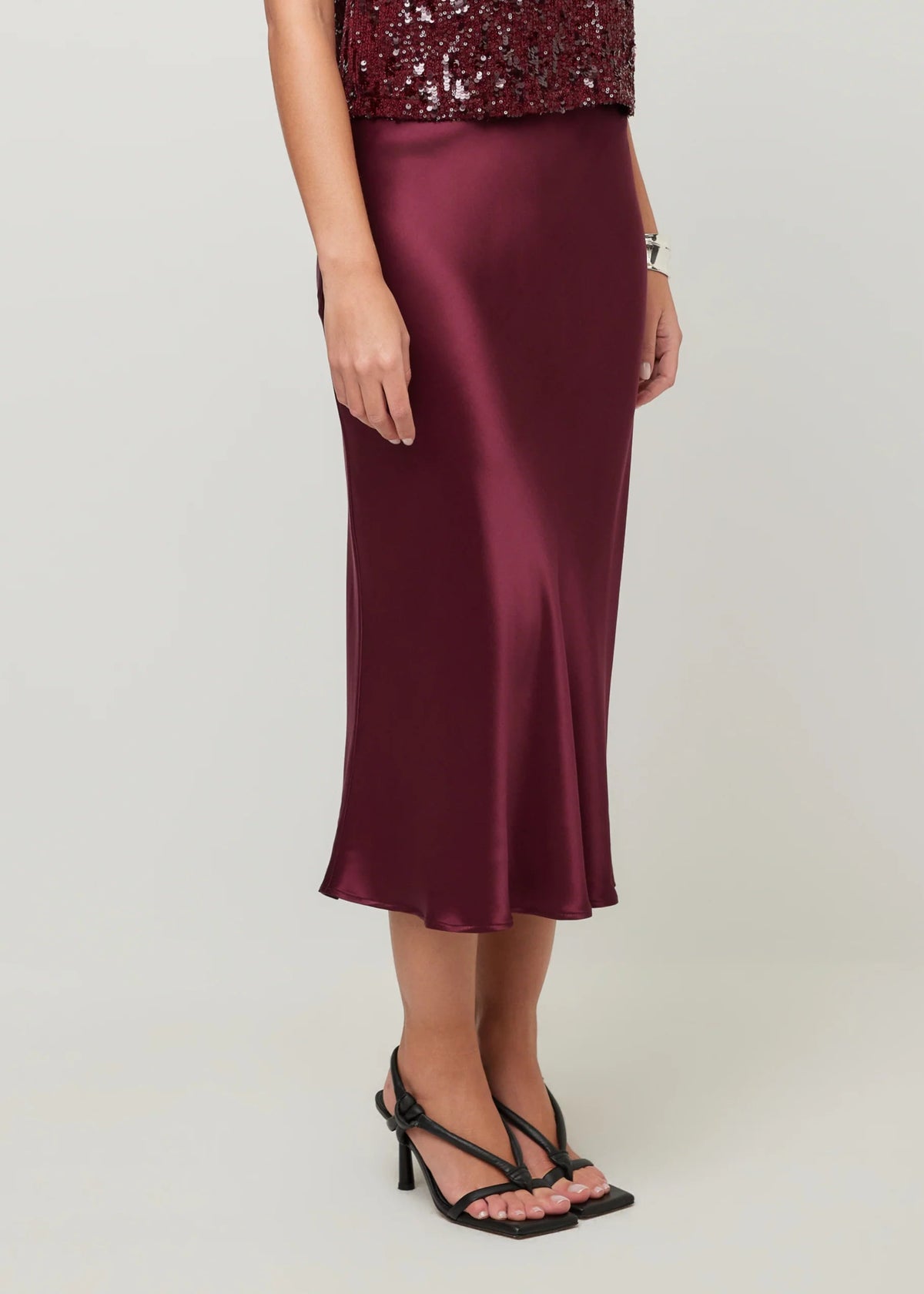 A woman wearing a satin midi skirt in deep burgundy, paired with black strappy high-heeled sandals and a sequined top, standing against a light gray studio background.