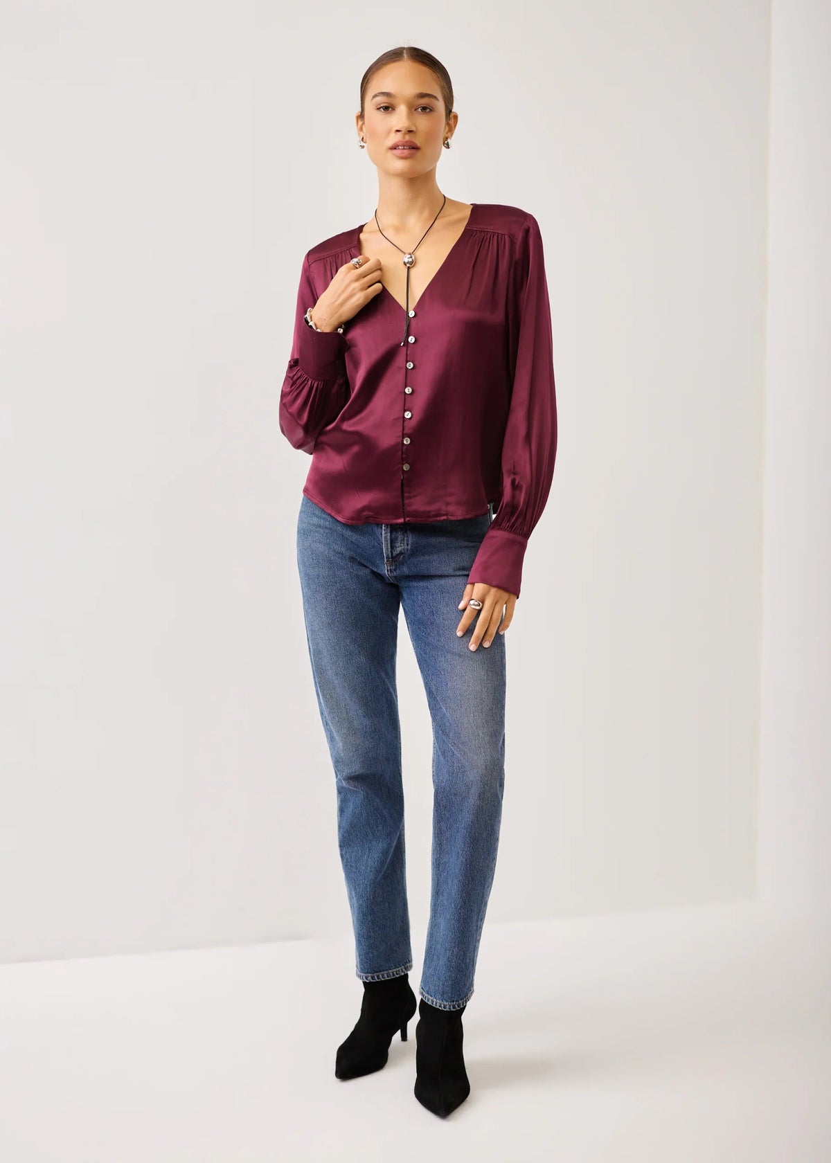 A woman modeling a long-sleeve satin blouse in deep burgundy with a button-down front, paired with blue jeans and black ankle boots, standing against a plain white studio background.