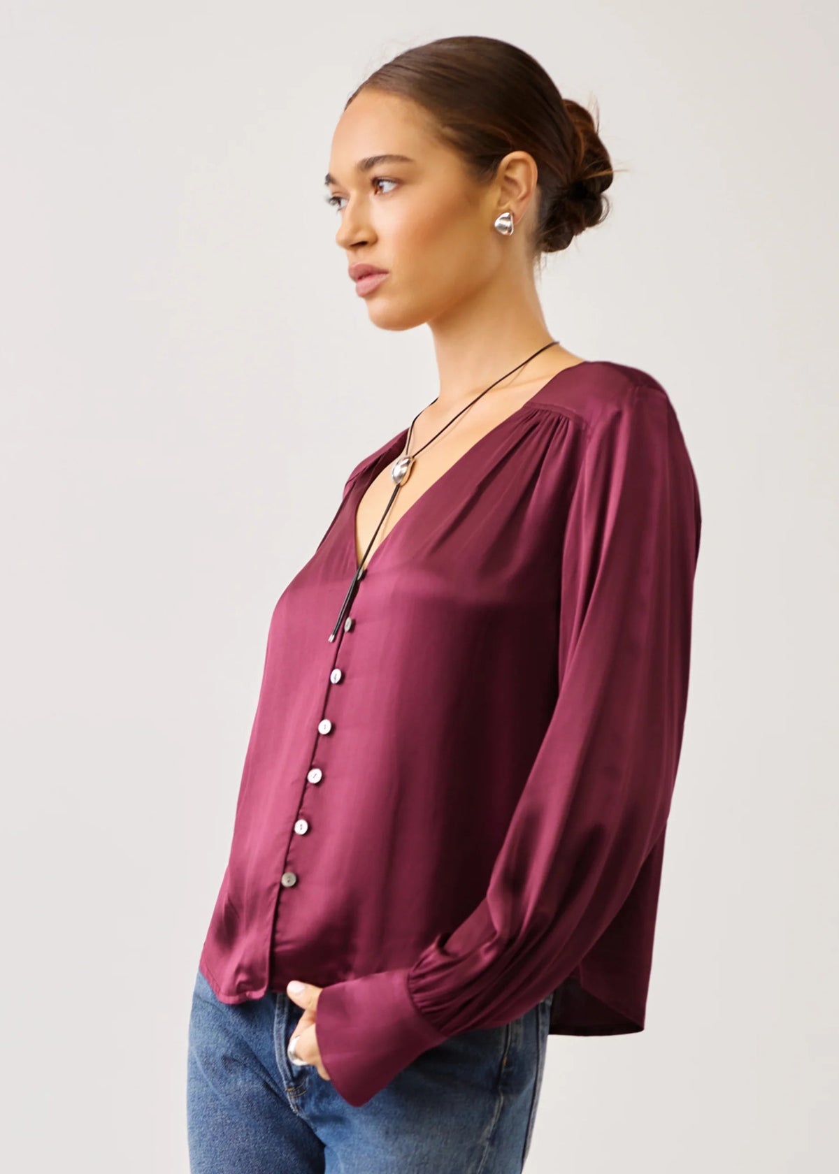 A woman wearing a long-sleeve satin blouse in deep burgundy with a button-down front and gathered neckline, paired with blue jeans, photographed against a light gray studio background.