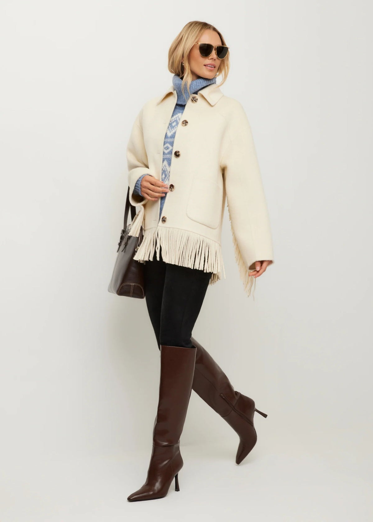 A woman models a cream-colored fringed jacket over a blue patterned top, black leggings, and brown knee-high boots, against a plain light gray studio background.